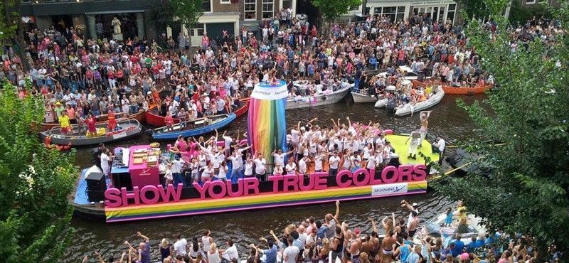 A canal filled with boats during a Pride parade, featuring a large float decorated with rainbows and