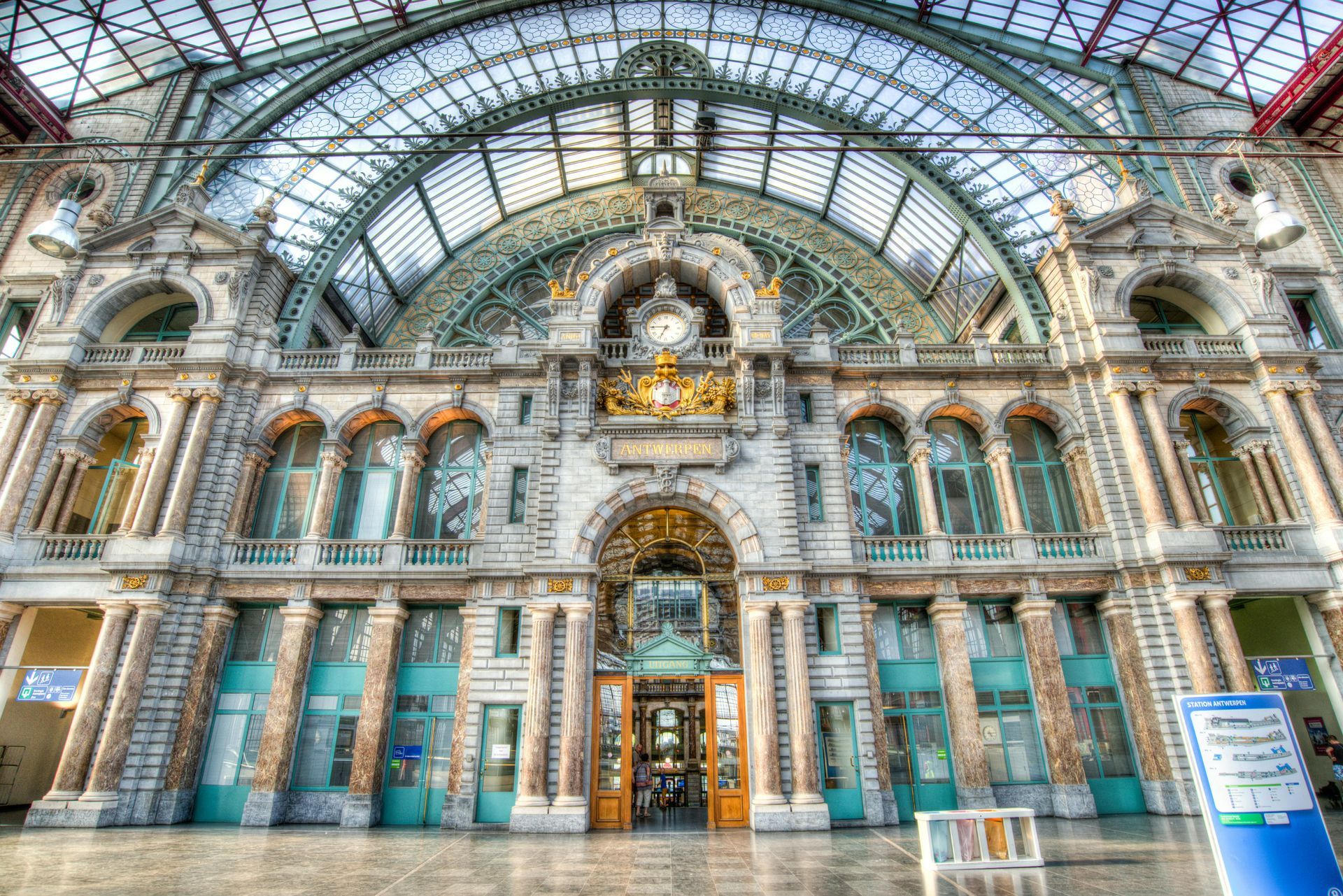 The ornate, vaulted interior of Antwerp Central Station, featuring a large glass dome, stone arches, and a central clock.