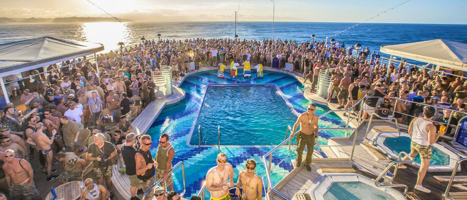 Large group of people gathered around a pool on a boat, ocean in background. People in and out of the water.