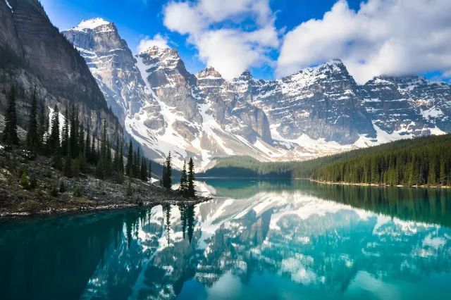 Turquoise lake reflects snow-capped mountains and evergreen trees under a bright blue sky. Banff National Park