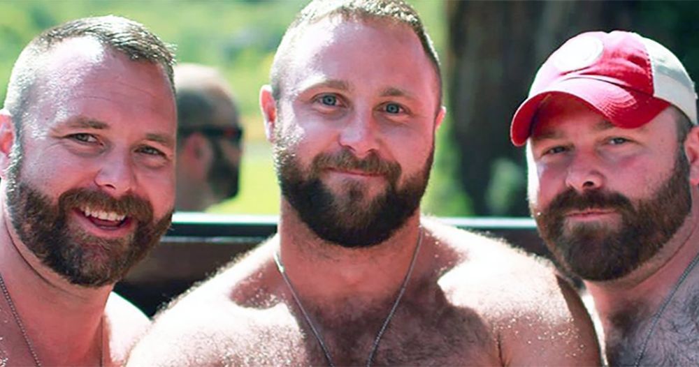 Three bearded men smiling together outdoors, one wearing a red and white baseball cap.