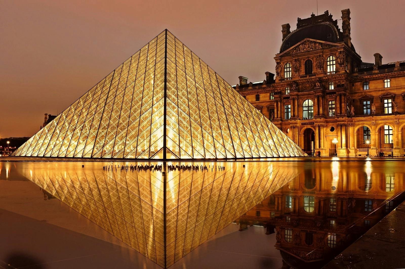 Louvre Pyramid at night, golden glass structure reflecting in water, beside illuminated classic building in Paris.