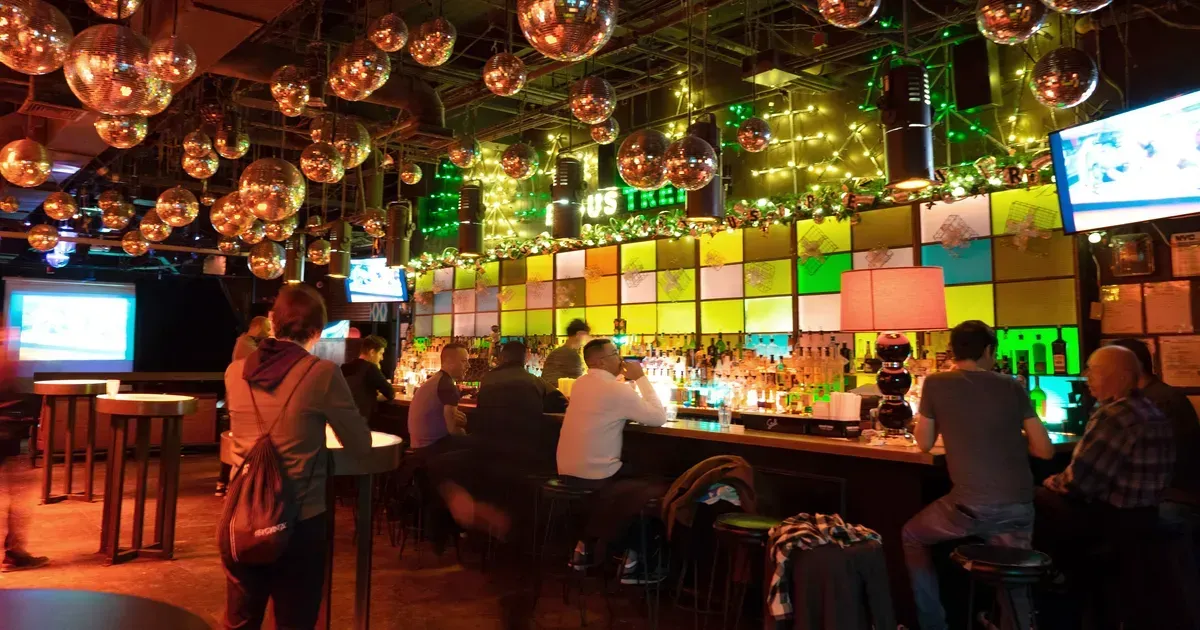 Bar interior with disco balls, patrons seated at the bar, colorful wall backdrop, and illuminated elements.