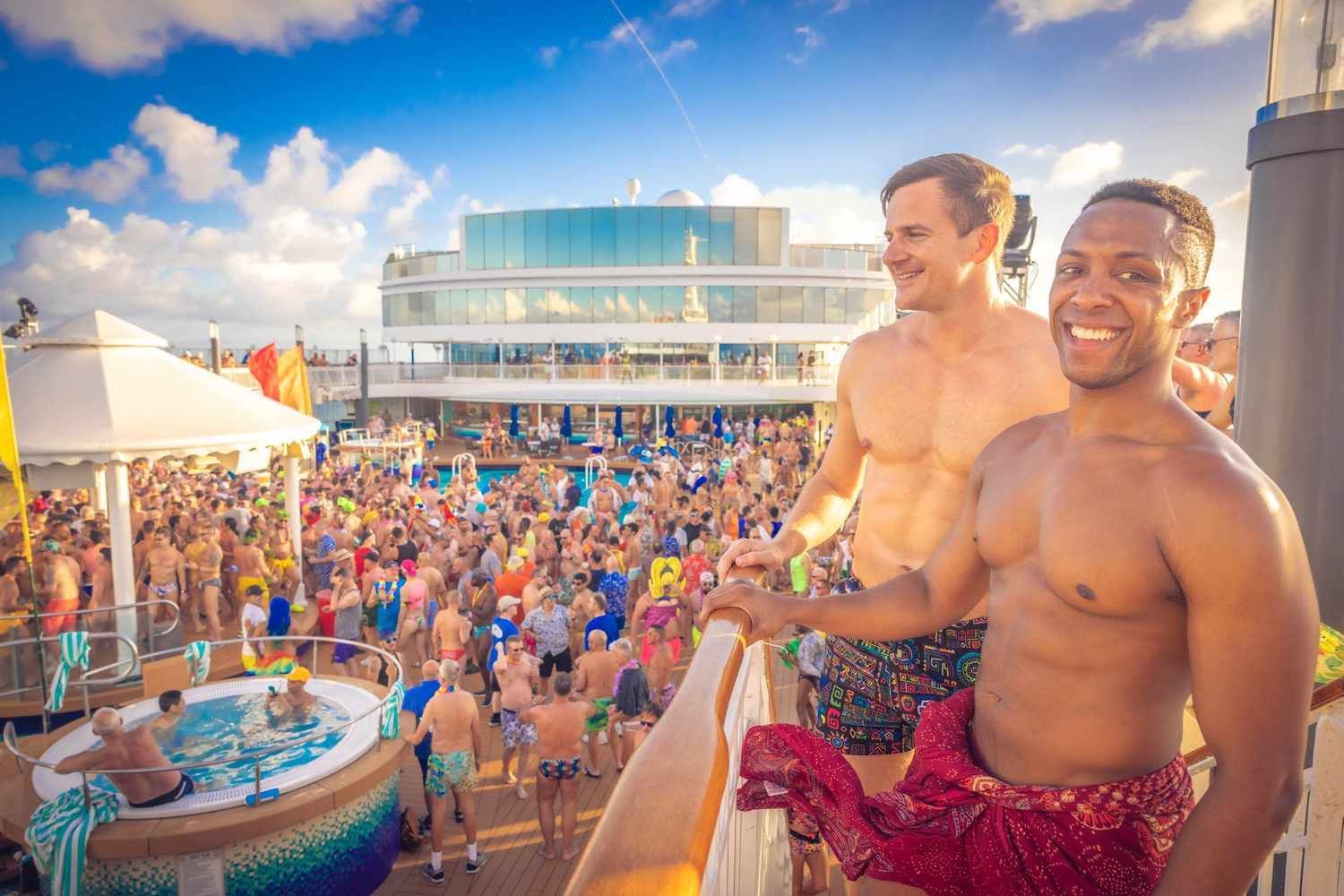 Two shirtless men stand on a cruise ship deck overlooking a crowded pool party with a hot tub and bright blue sky.