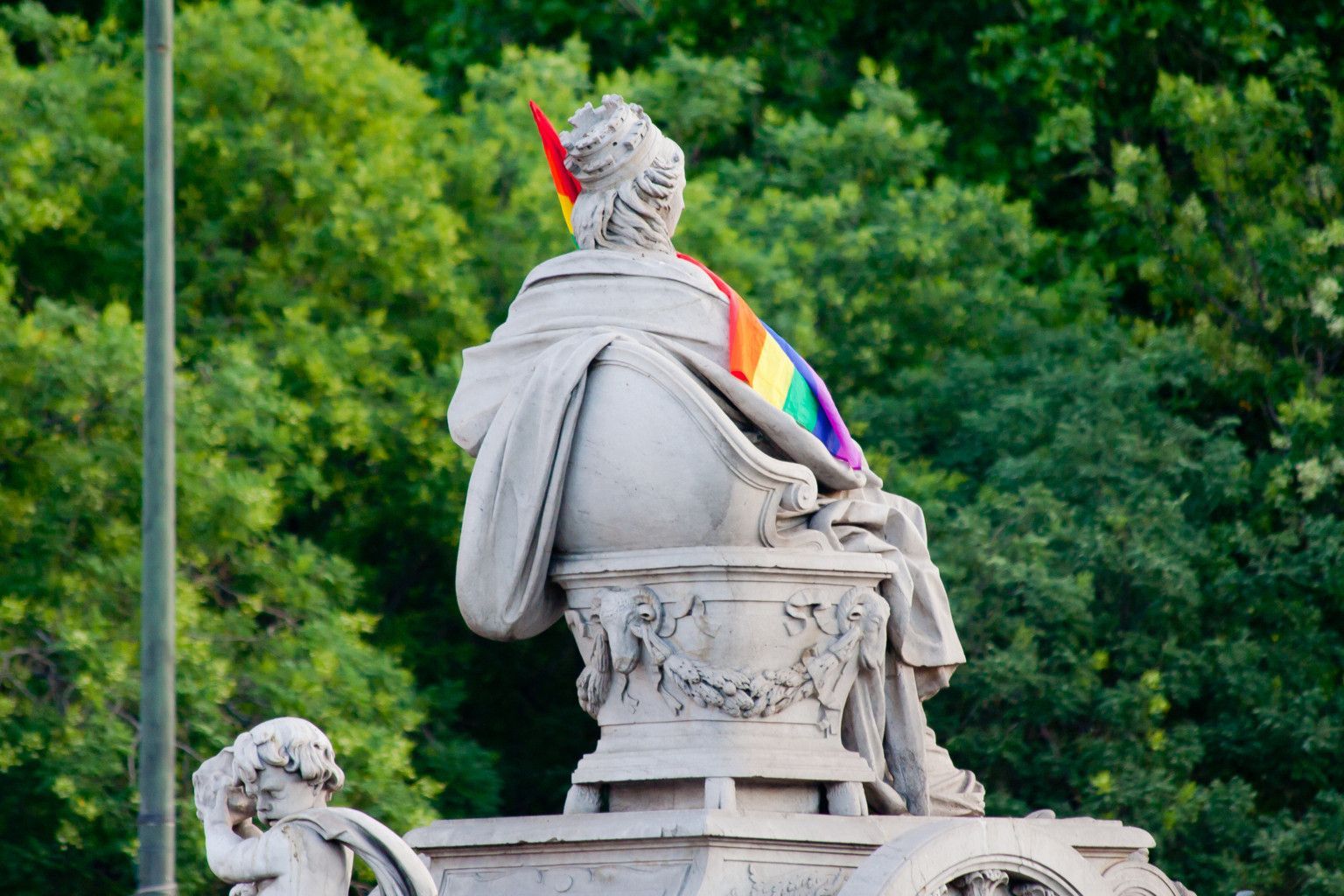 Statue draped in rainbow pride flag, set against green foliage.