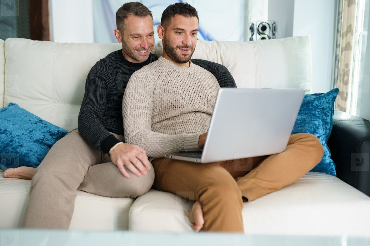 Two people sit on a couch using a laptop, with blue pillows in the background.