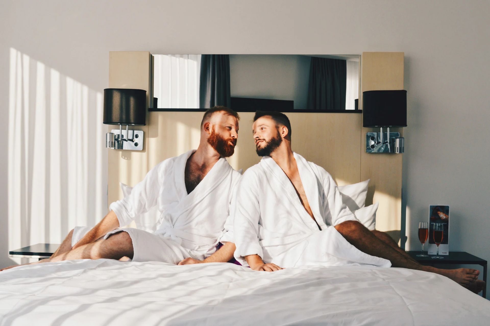 Two men in white robes looking at each other on a bed in a hotel room, sunlight streaming in.