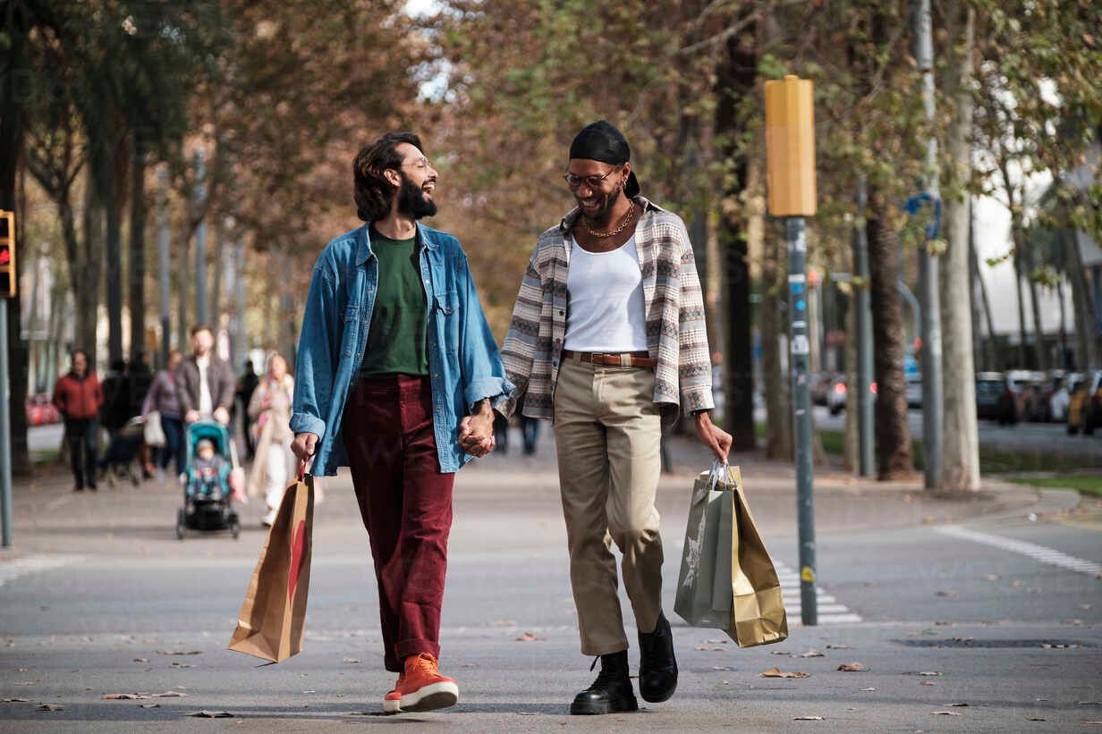 Two people walking down a tree-lined street, holding hands and shopping bags, smiling at each other.