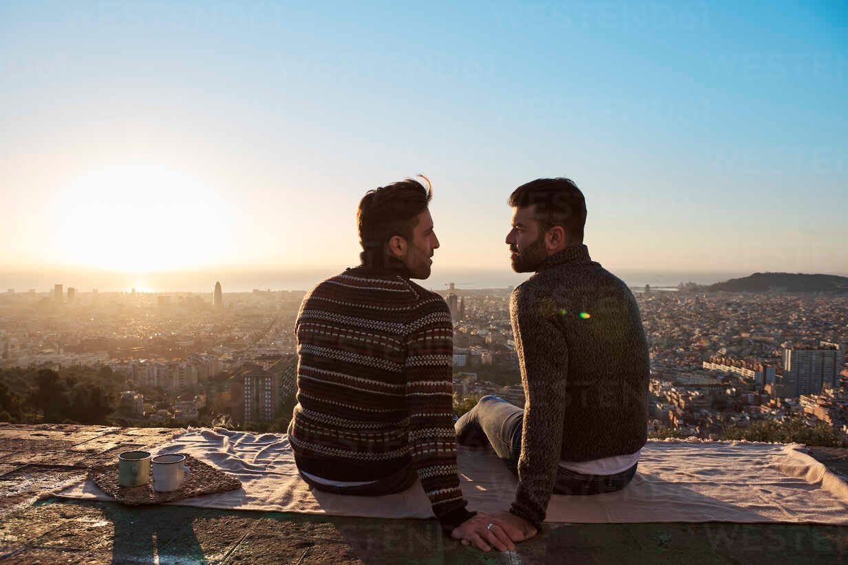 Two people sit on a ledge overlooking a city at sunset, facing each other with warm light behind them.