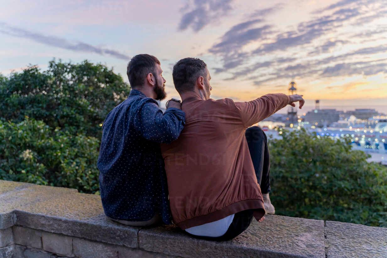 Two people sitting on a wall at sunset, one pointing toward a city view while the other looks on.