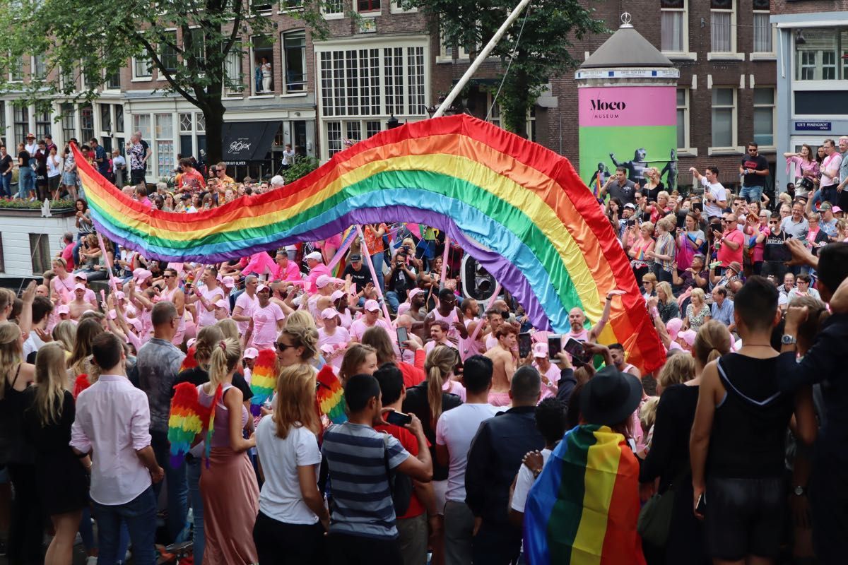 A large rainbow flag held high over a vibrant, crowded outdoor Pride parade in a city with historical buildings.