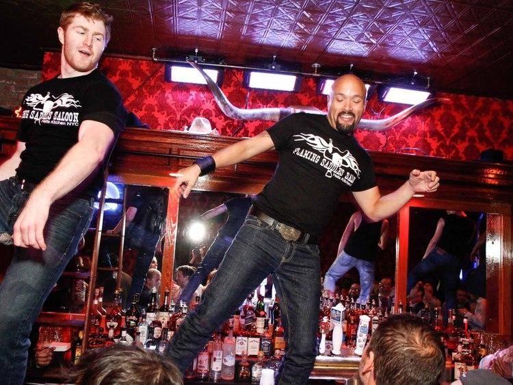 Two men dancing on a bar, wearing black t-shirts and jeans in a dimly lit bar with a decorative ceiling.