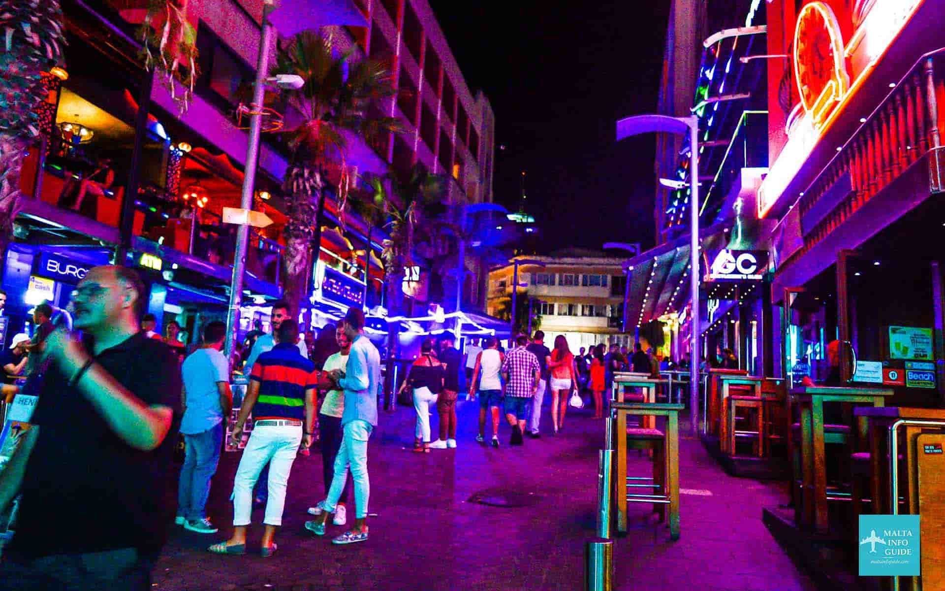 Neon-lit city street at night with pedestrians, bars, and colorful signs reflected on wet pavement