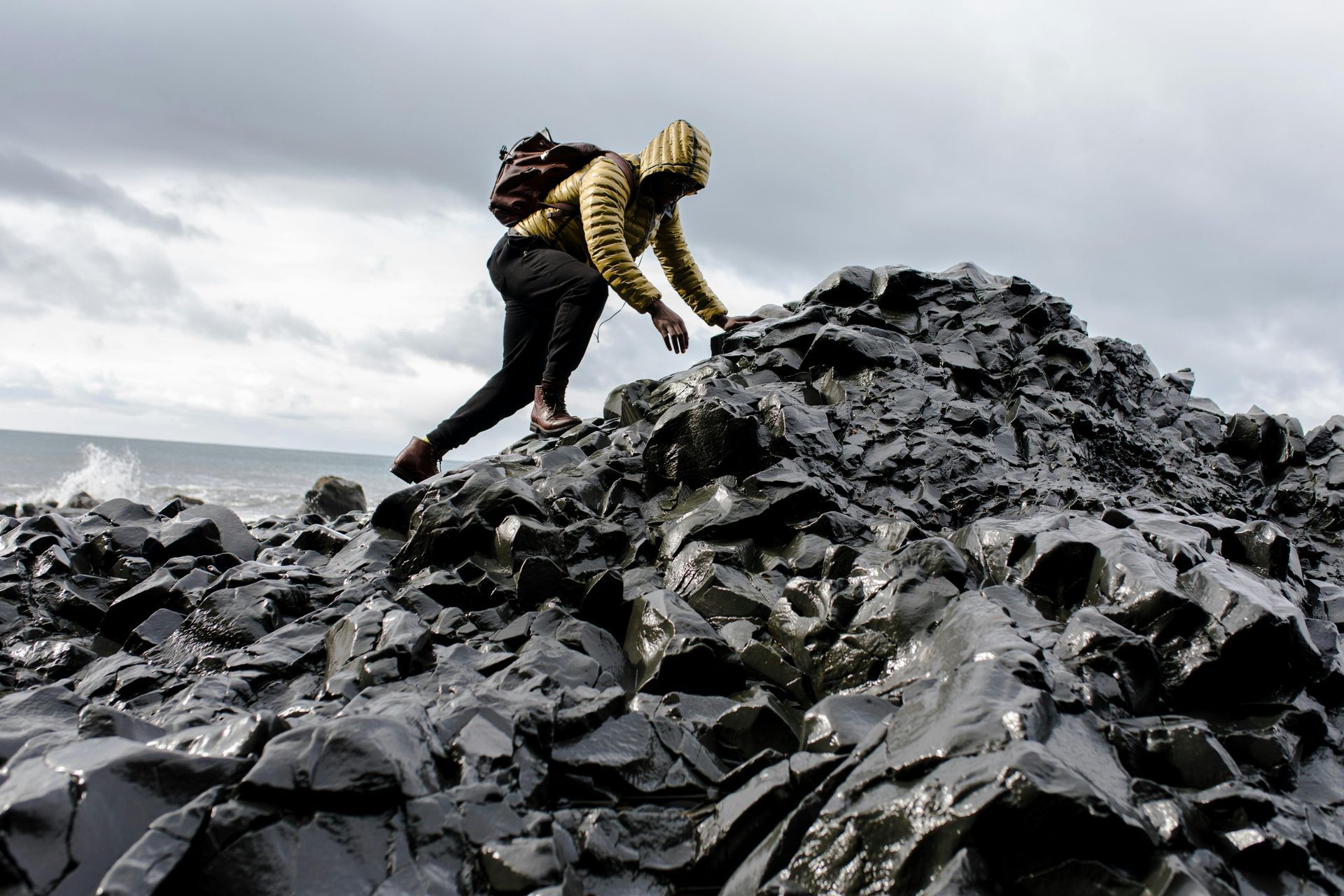 Person in a yellow hoodie climbs dark, wet rocks on a cloudy day; ocean in the background.