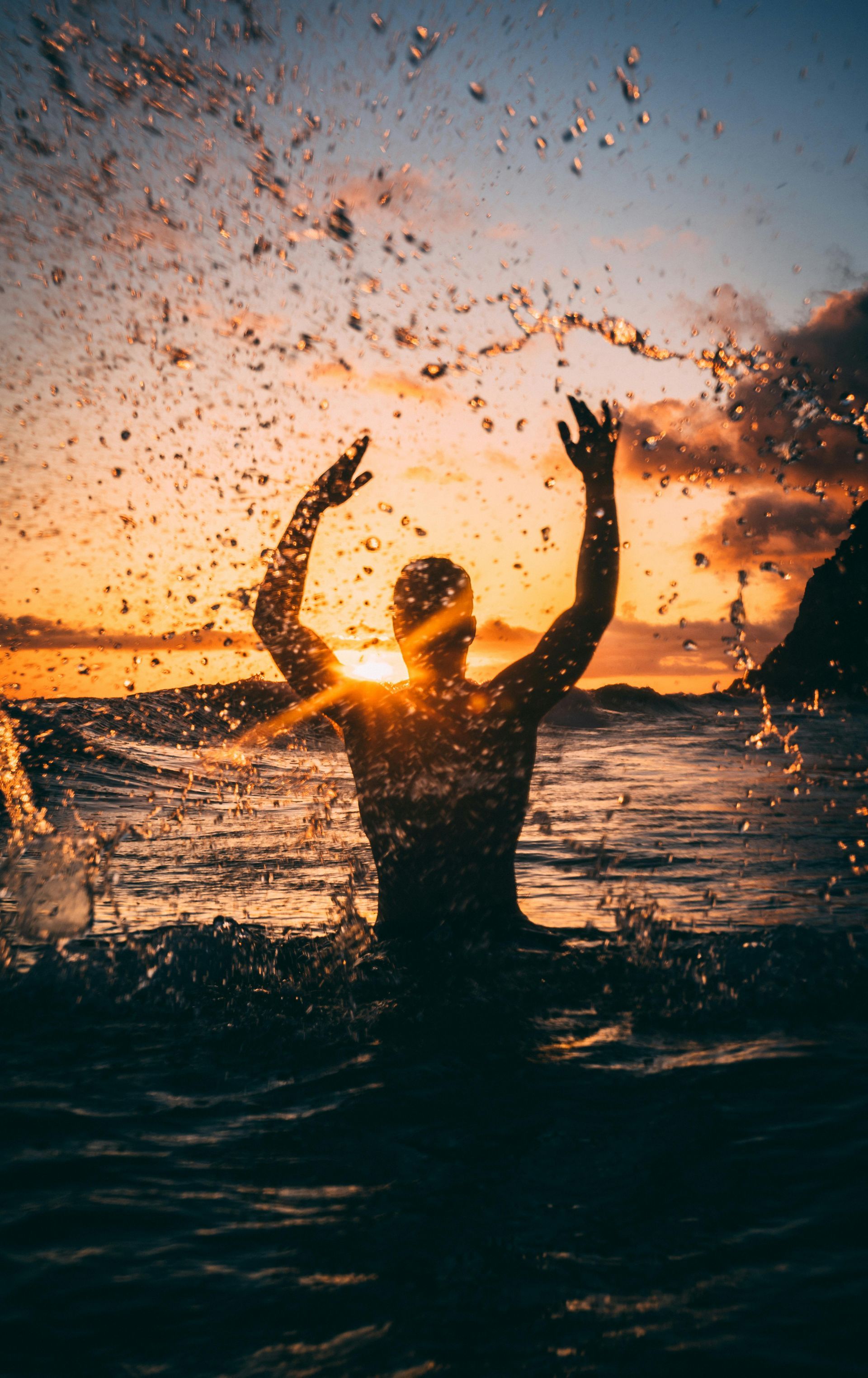 A person stands in ocean waves with arms raised as water splashes around them during a sunset.