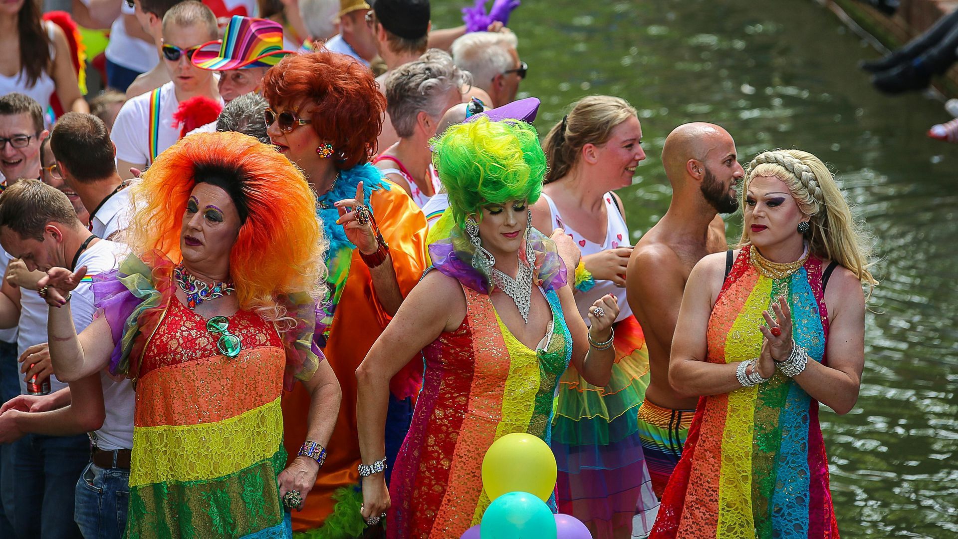 People in bright, rainbow-themed costumes and vibrant wigs celebrate at a daytime pride parade near a canal.