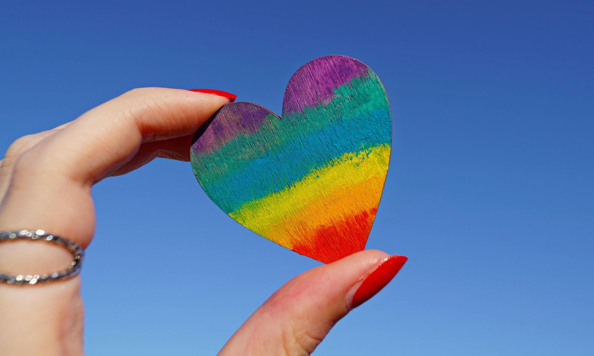 Hand holding a rainbow-colored heart against a bright blue sky, symbolizing LGBTQ+ pride.