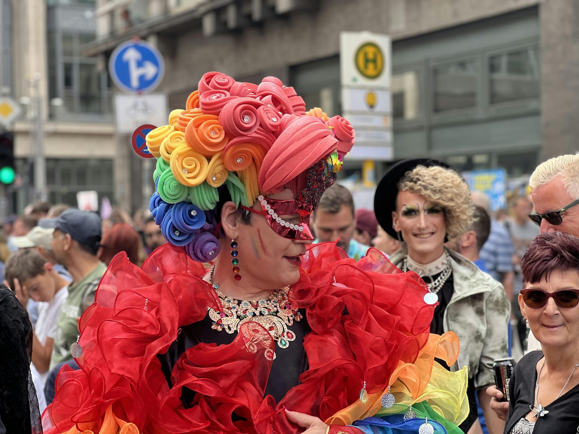 Person in elaborate rainbow costume at a Pride parade, red ruffled dress, colorful headdress, smiling, crowd in the background.