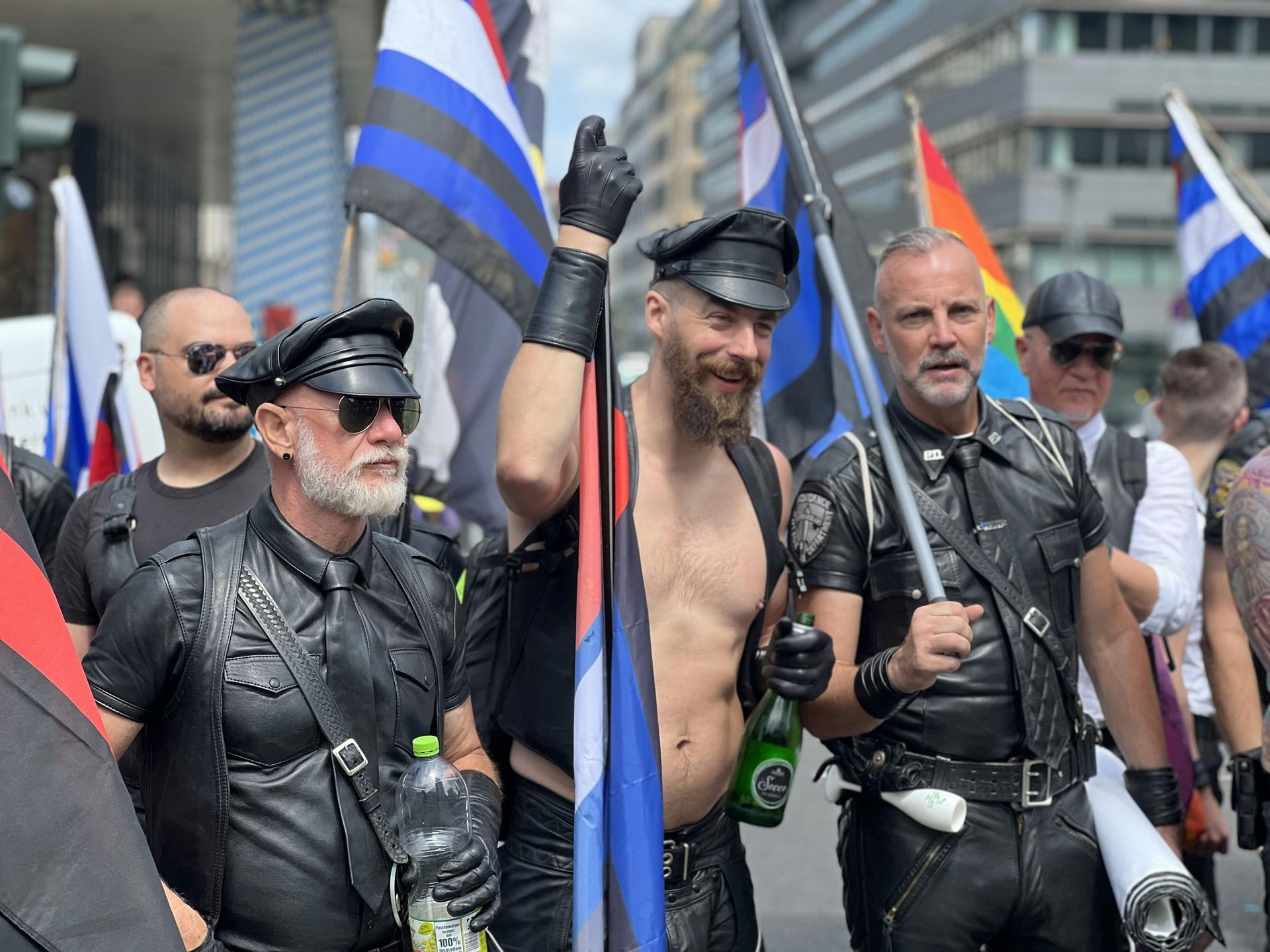 Men in leather gear and hats march at a Pride parade, holding flags.
