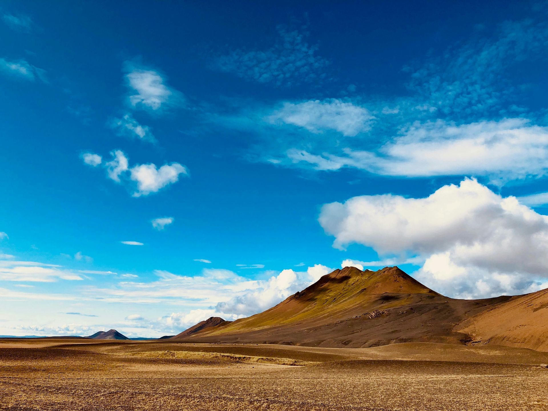 Vast arid landscape under a bright blue sky with fluffy white clouds, featuring a brown mountain.