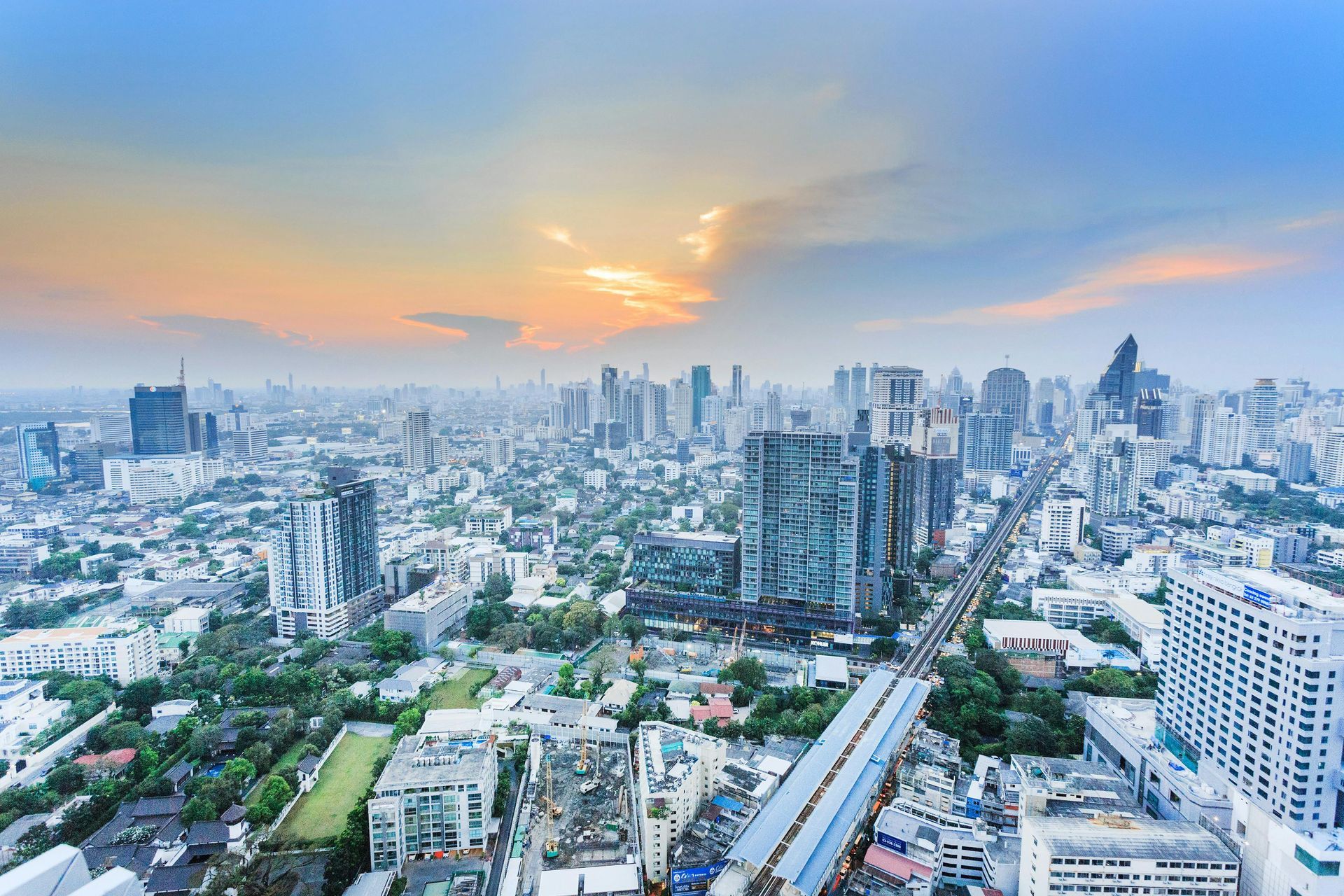 City skyline under a blue and orange sky, with numerous tall buildings and a train line.Bangkok