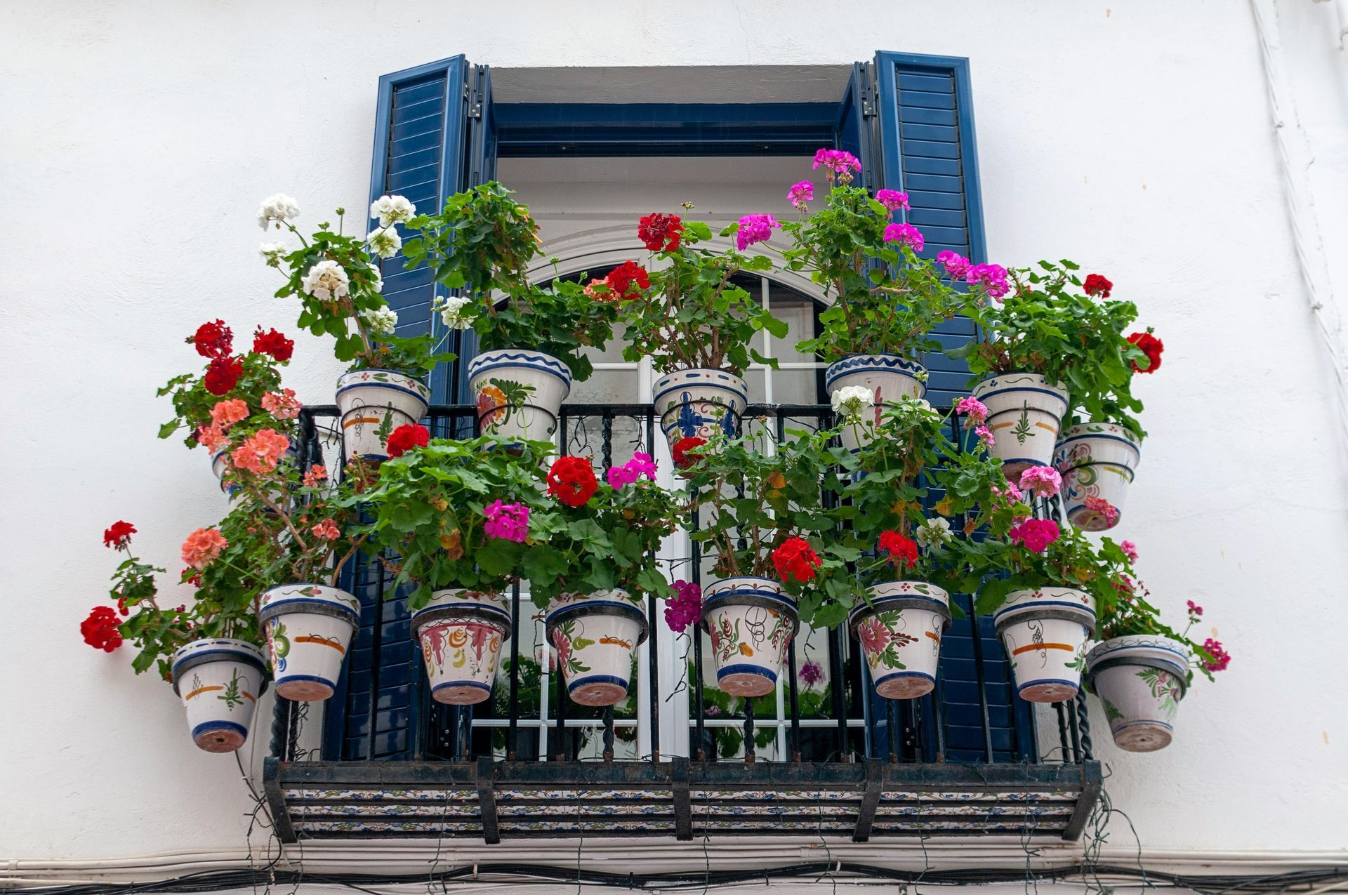 Balcony with colorful flowers in ceramic pots, against blue shutters and white building facade.