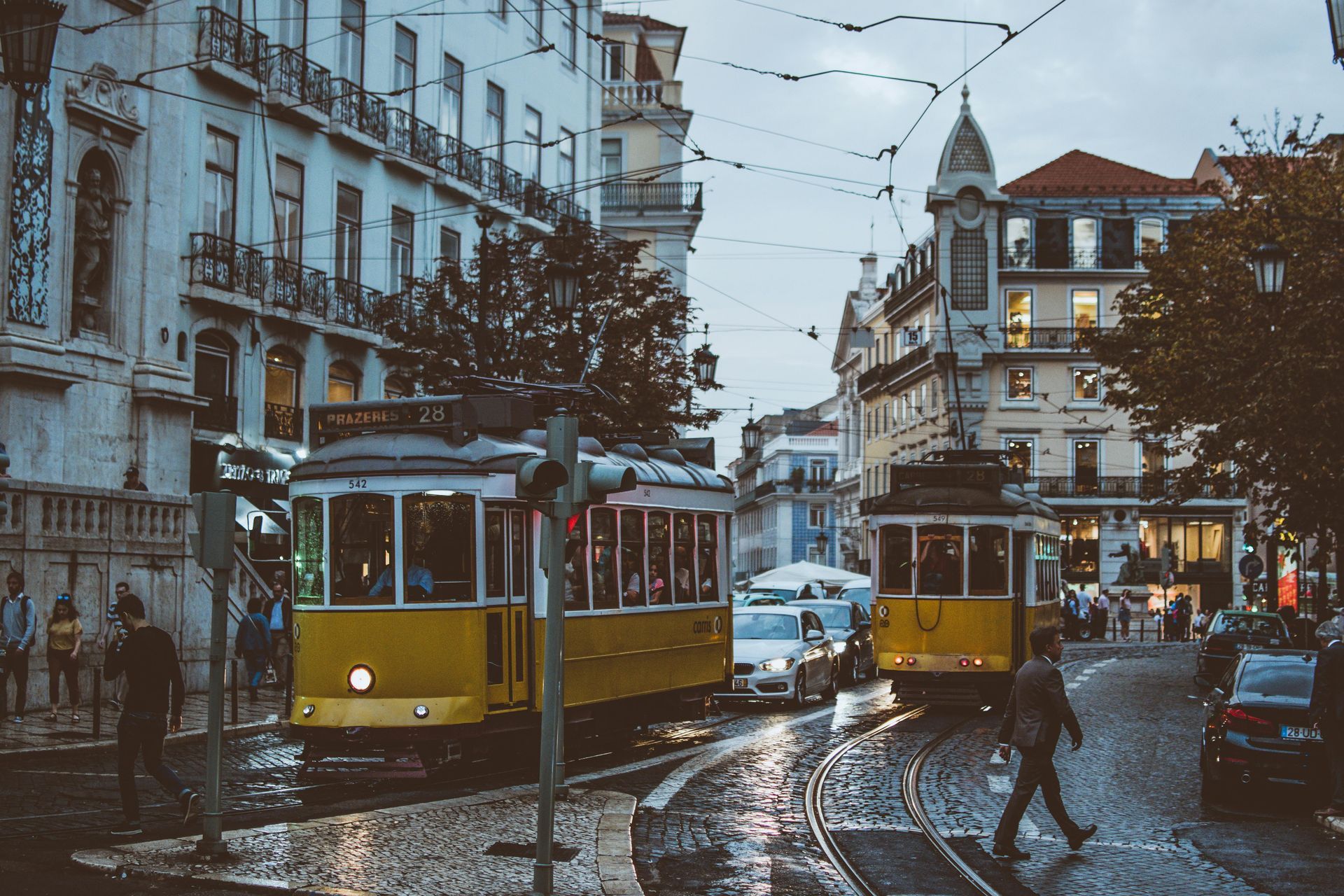 Yellow trams on a wet street in Lisbon, Portugal. Buildings line the road, and pedestrians cross. Lisbon