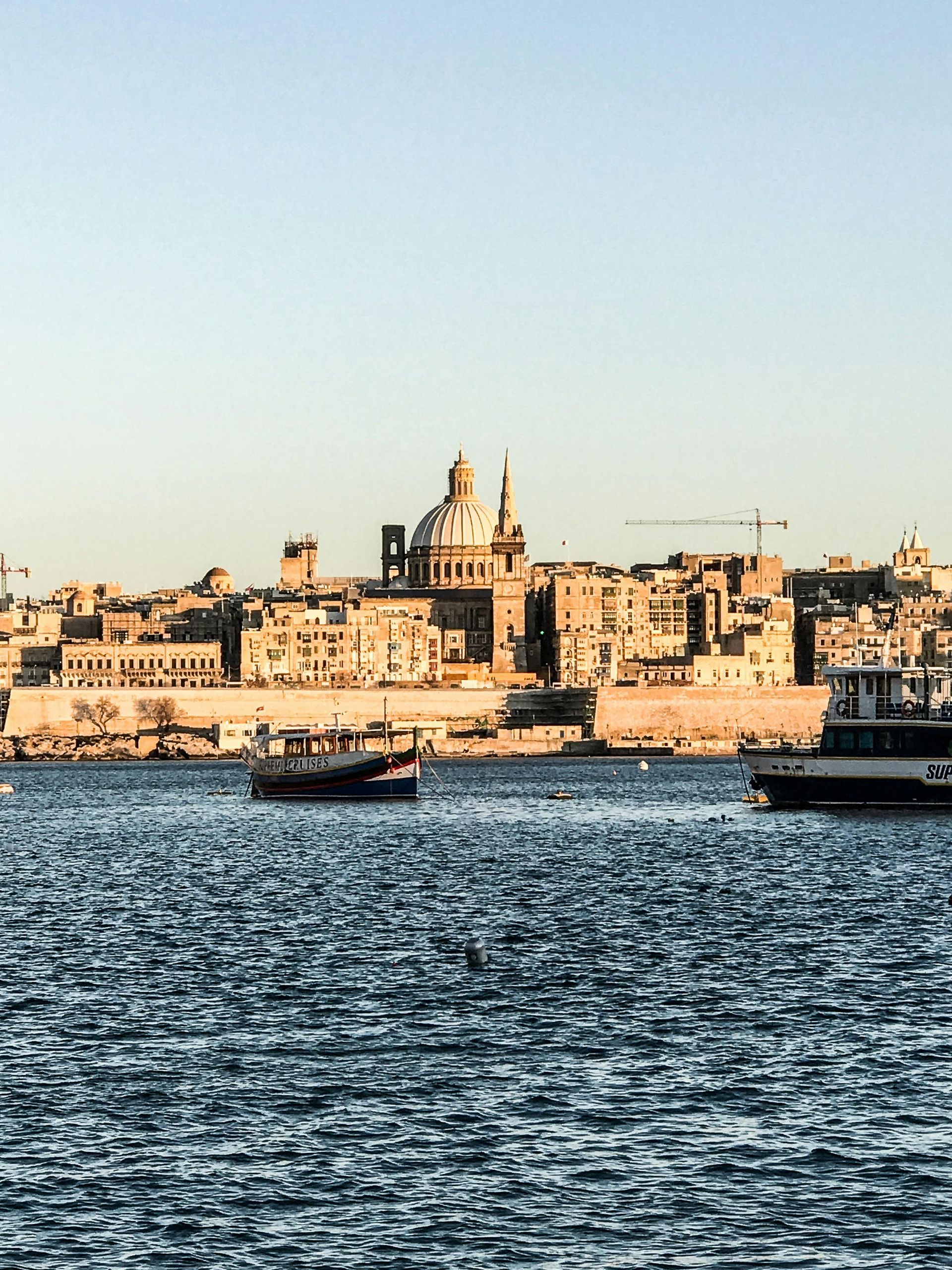 City skyline across choppy blue water at sunset, with historic buildings and boats in the harbor