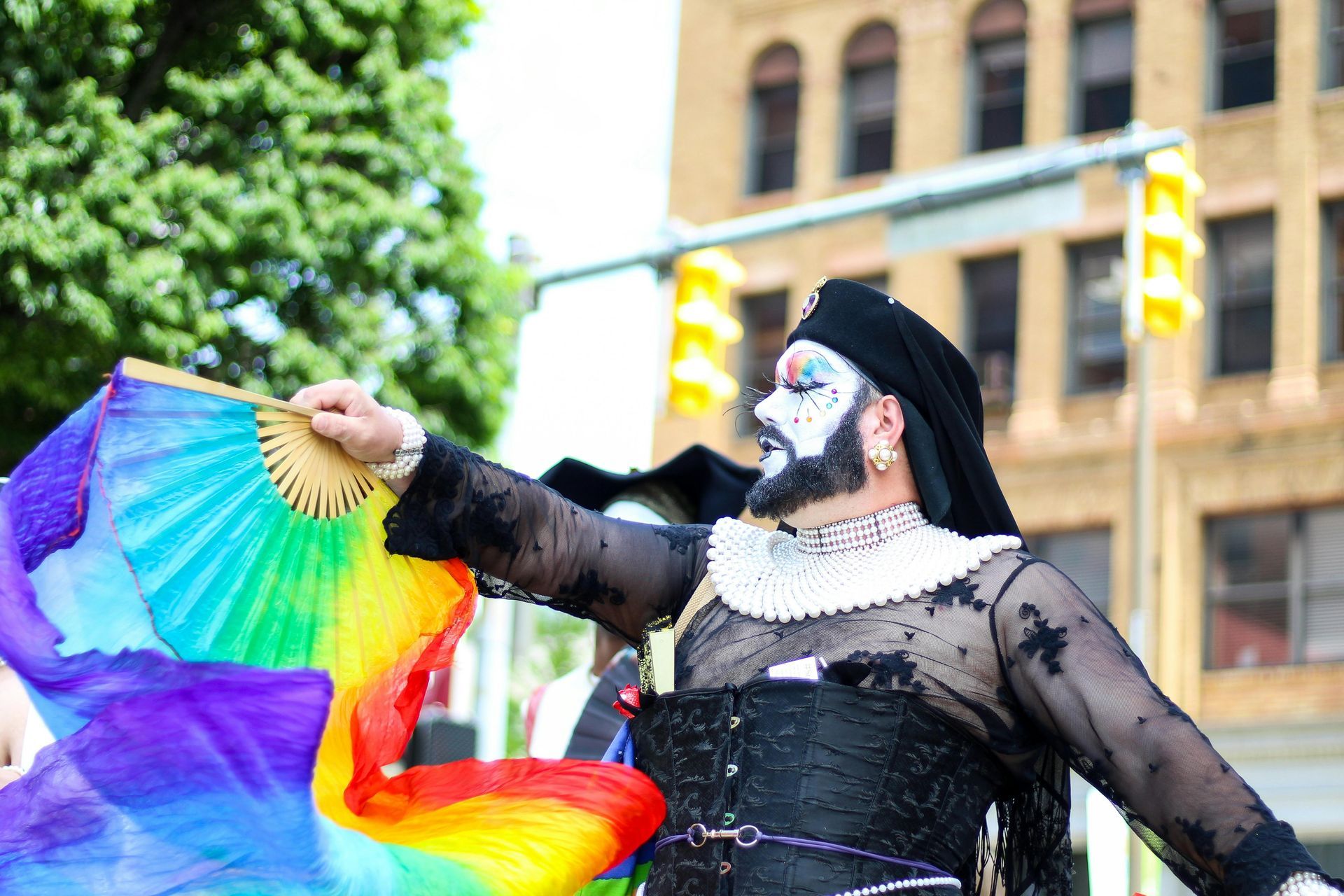Person in drag, holding rainbow fan, at a parade. Black outfit, makeup, buildings in background.