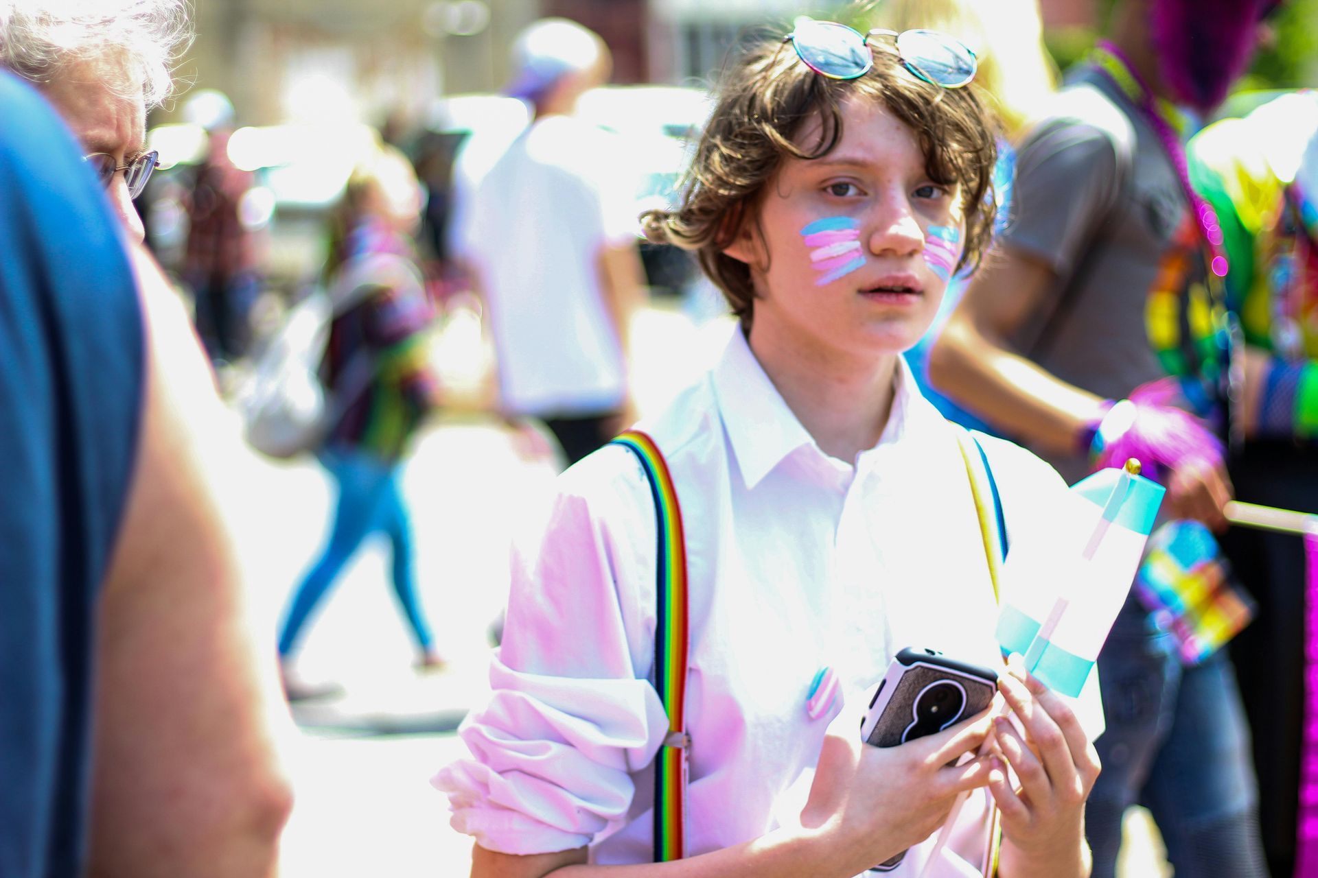 A person wearing rainbow suspenders and trans flag face paint holds a small flag at a pride parade.