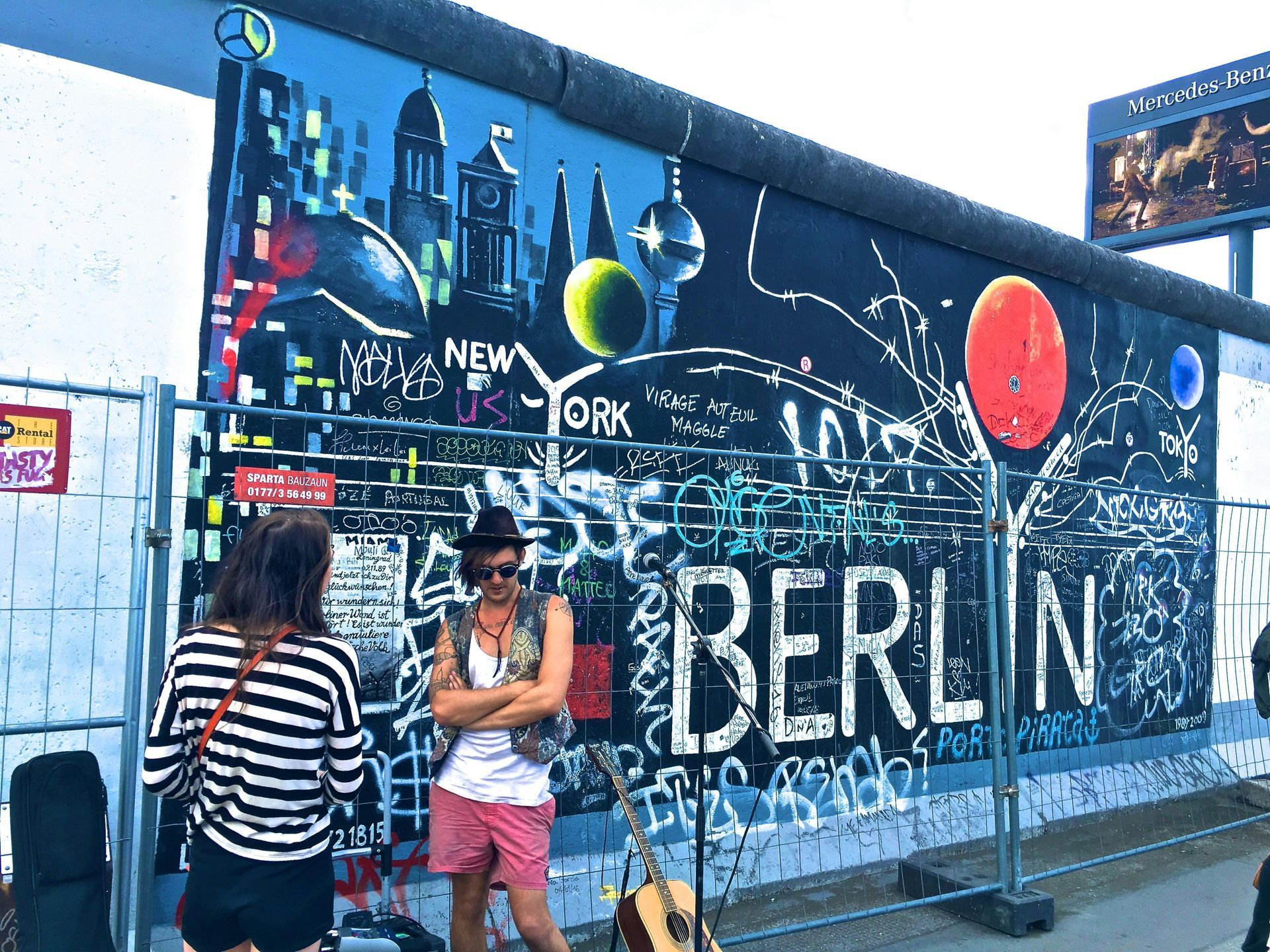 Two people stand before a colorful mural on the Berlin Wall. One with a guitar. Graffiti, cityscape.