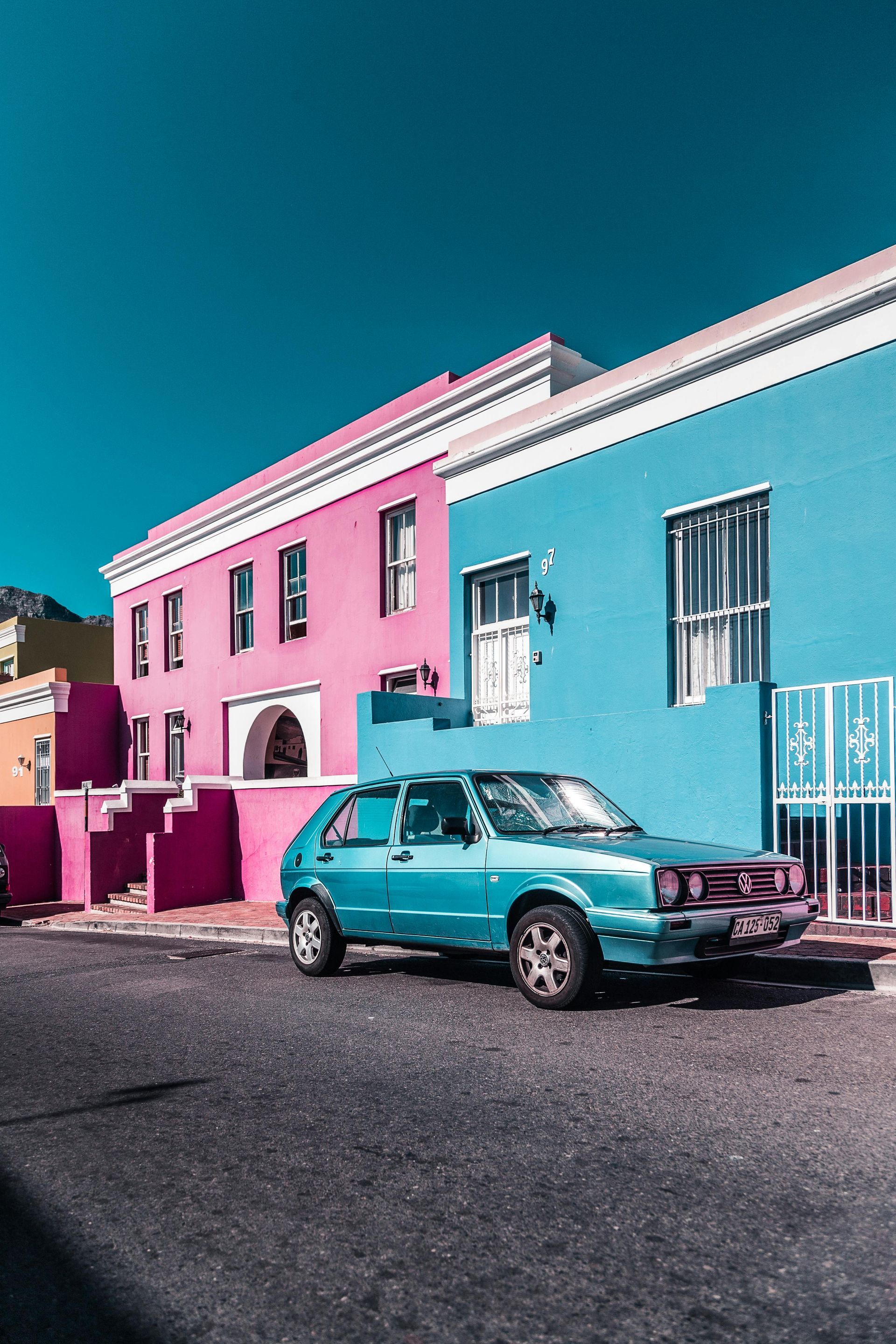 Colorful houses in pink, blue, and yellow line a street with a teal car parked in front; blue sky above. Gay City Breaks in Cape Town