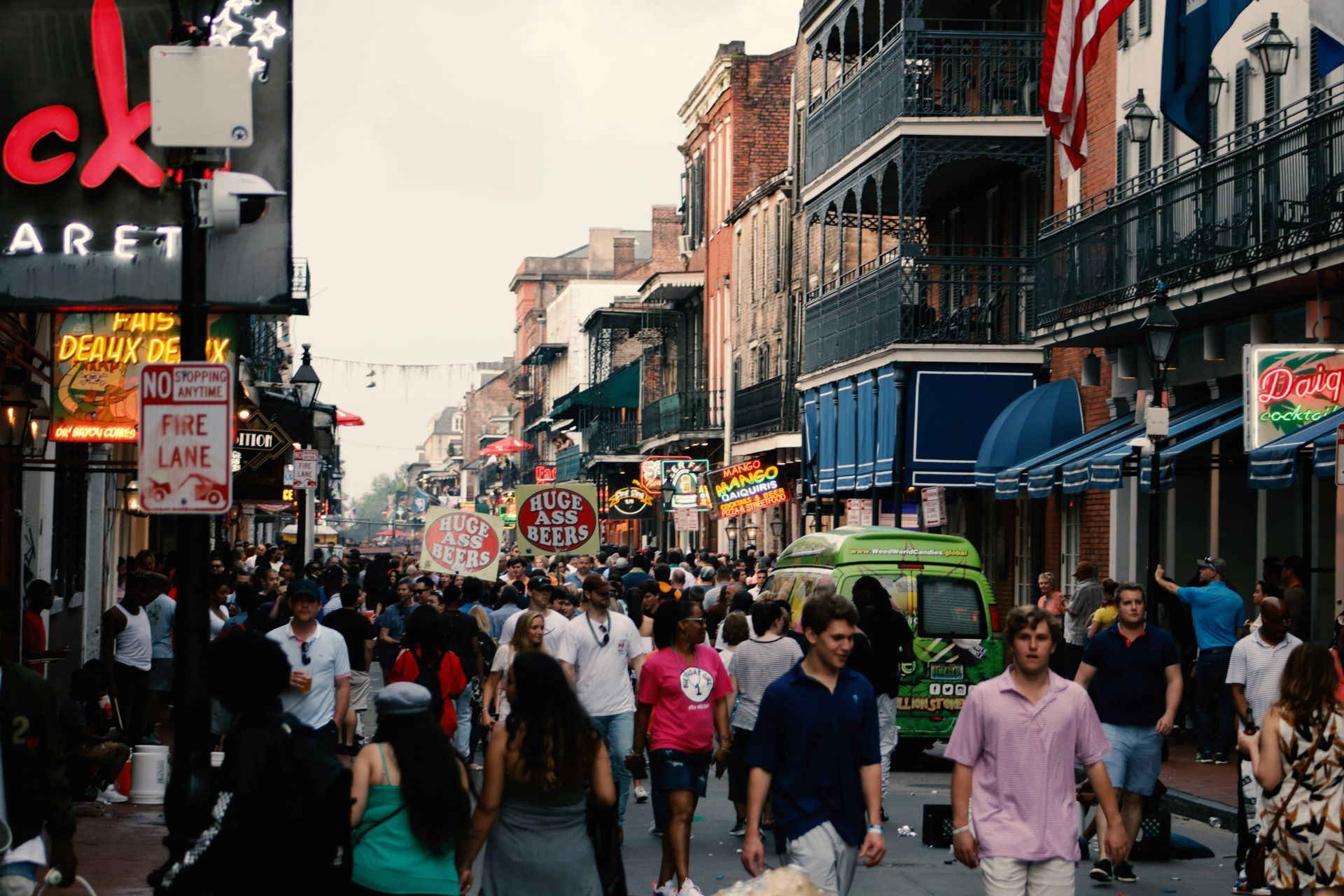 Crowd on a street in a city, buildings with balconies, shop signs, overcast sky.