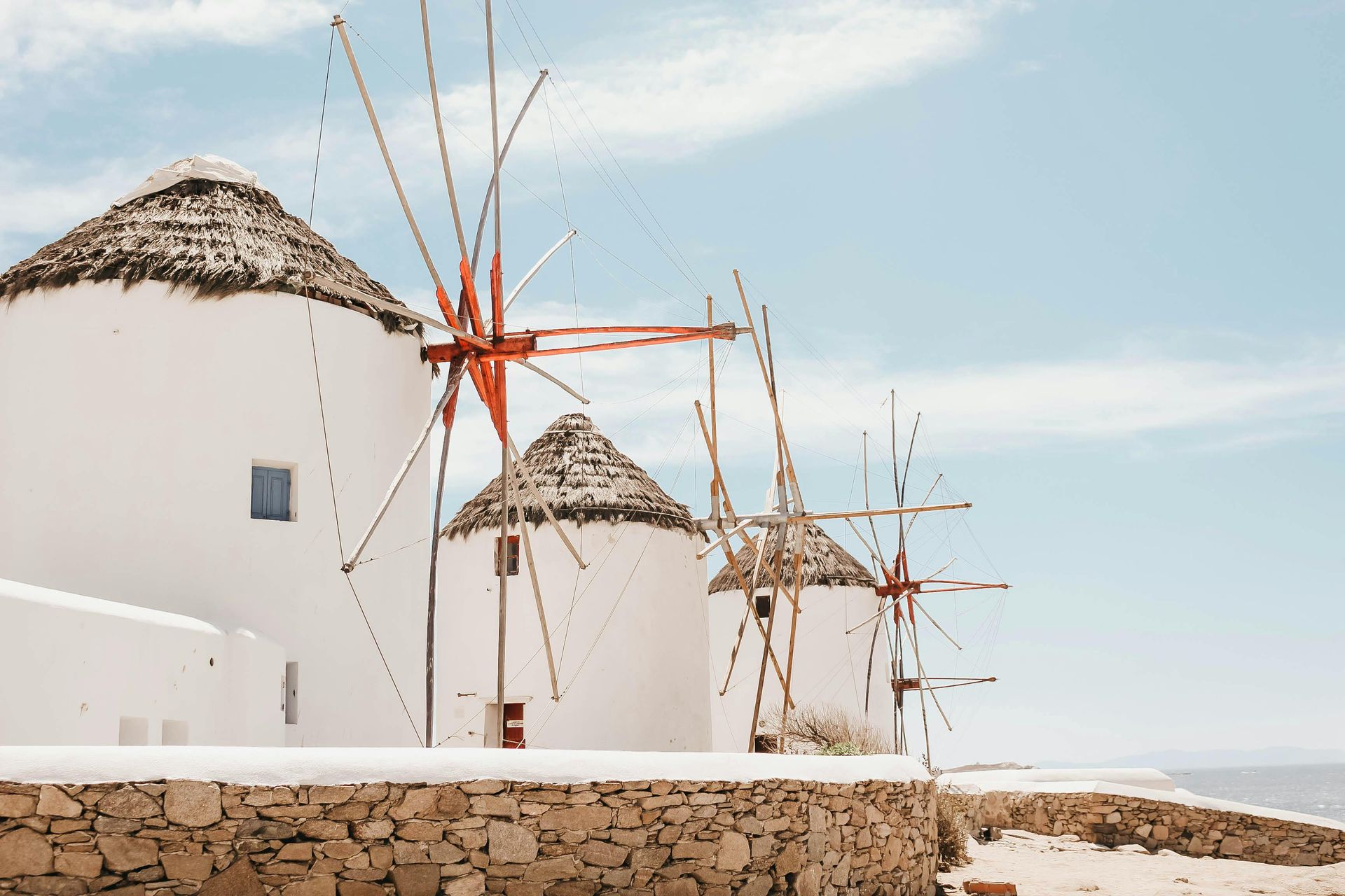 A row of iconic whitewashed windmills with thatched roofs on a stone wall against a bright sky on Mykonos, Greece.