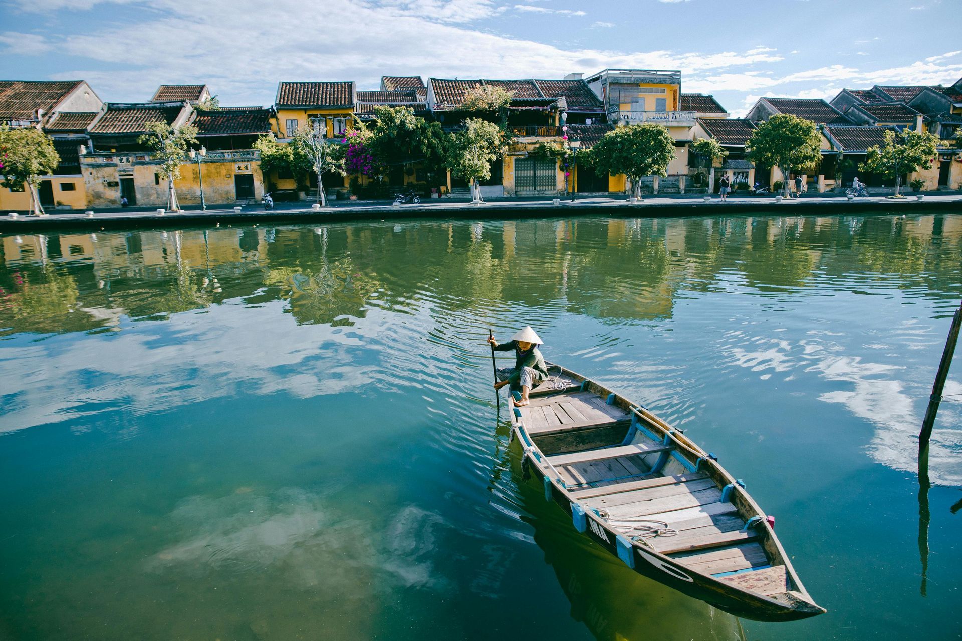 A person in a conical hat rows a small wooden boat on a calm river lined with traditional yellow buildings in Hoi An. Vietnam
