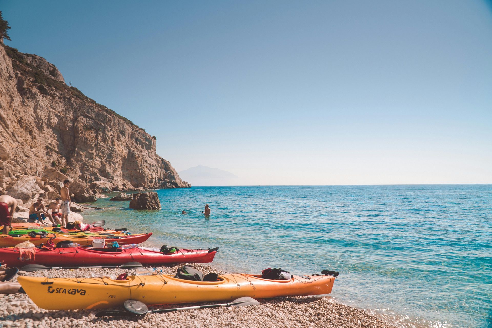 Kayaks on a pebbled beach, cliffside in background. People swimming in clear blue water under a sunny sky.