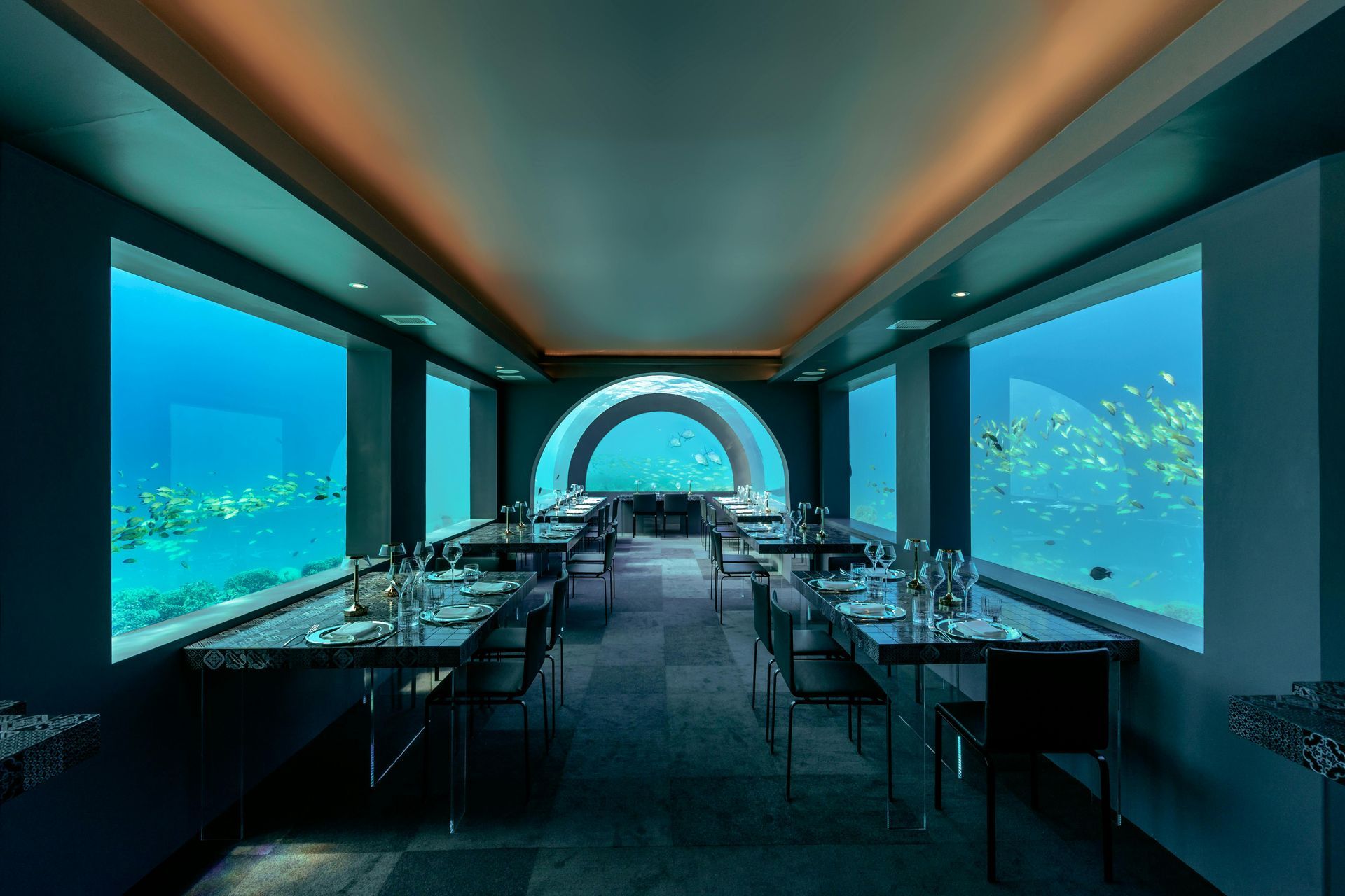 Underwater restaurant with tables set. Large windows provide ocean views, featuring coral and marine life.