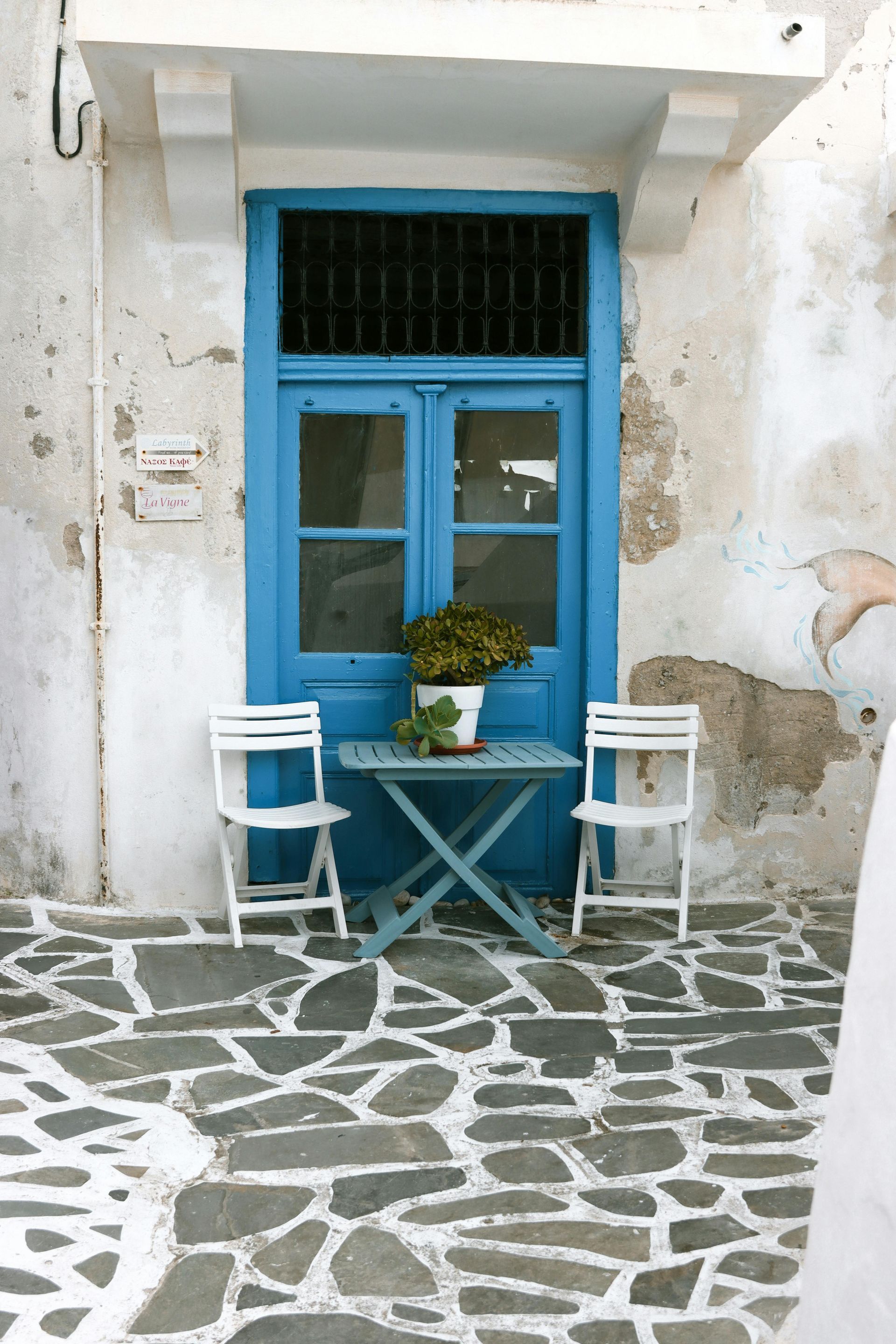 A bright blue door set in a textured, weathered white wall with two white chairs and a small table on a stone patio.