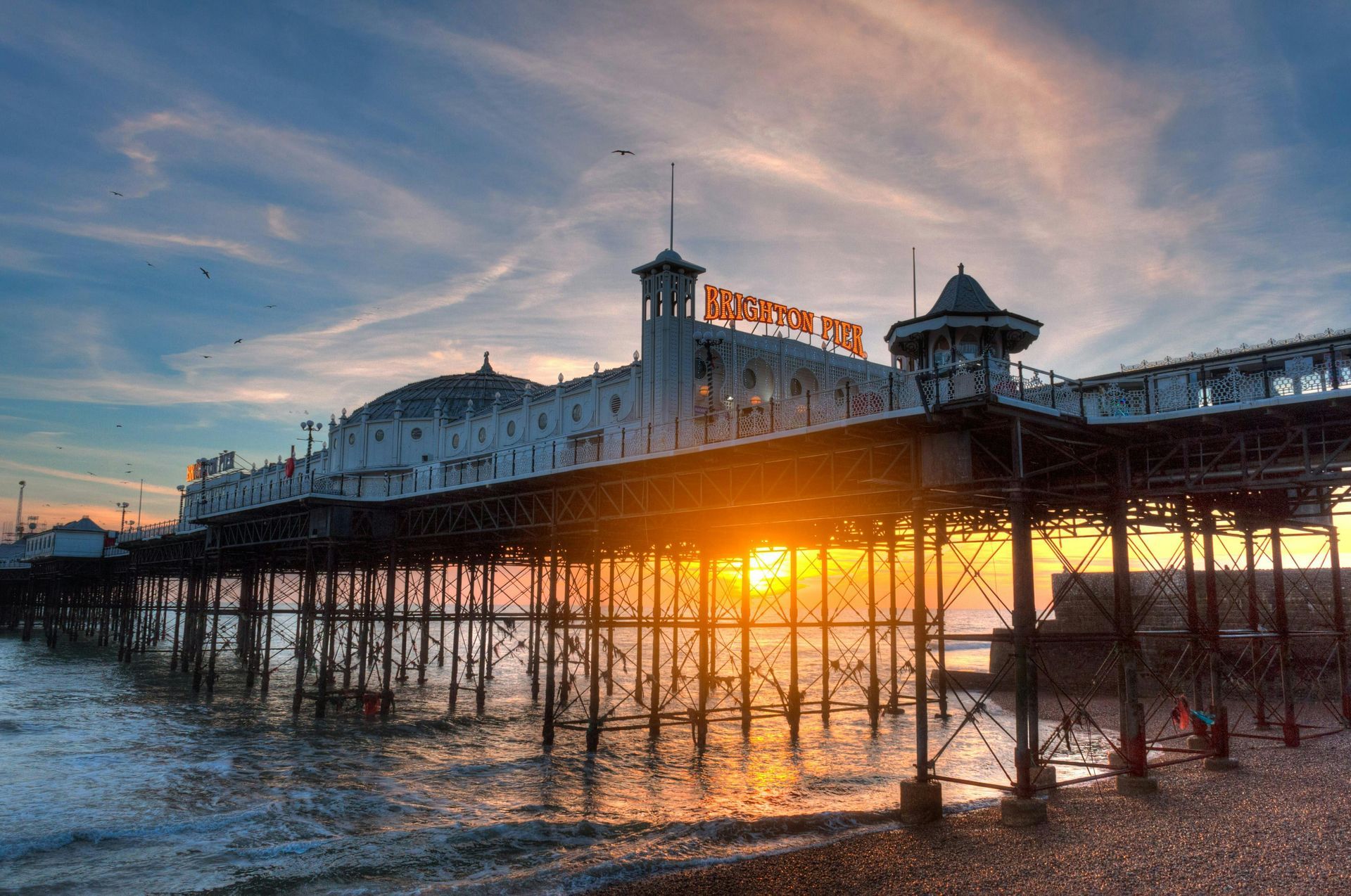 Sunset behind a seaside pier with a lit amusement sign and people silhouetted on the deck