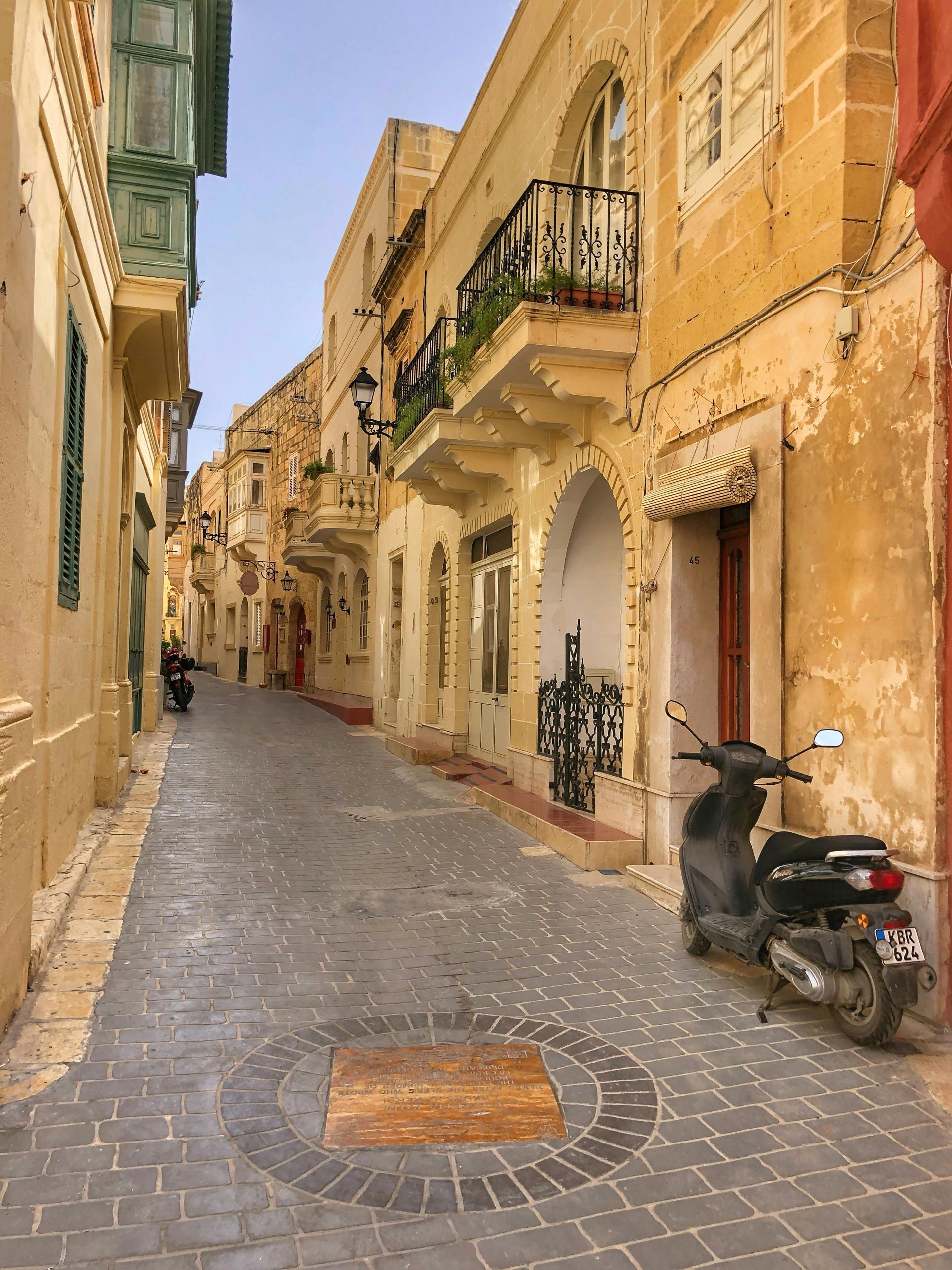 Narrow cobblestone street lined with old yellow buildings and balconies, with a parked scooter on the right.