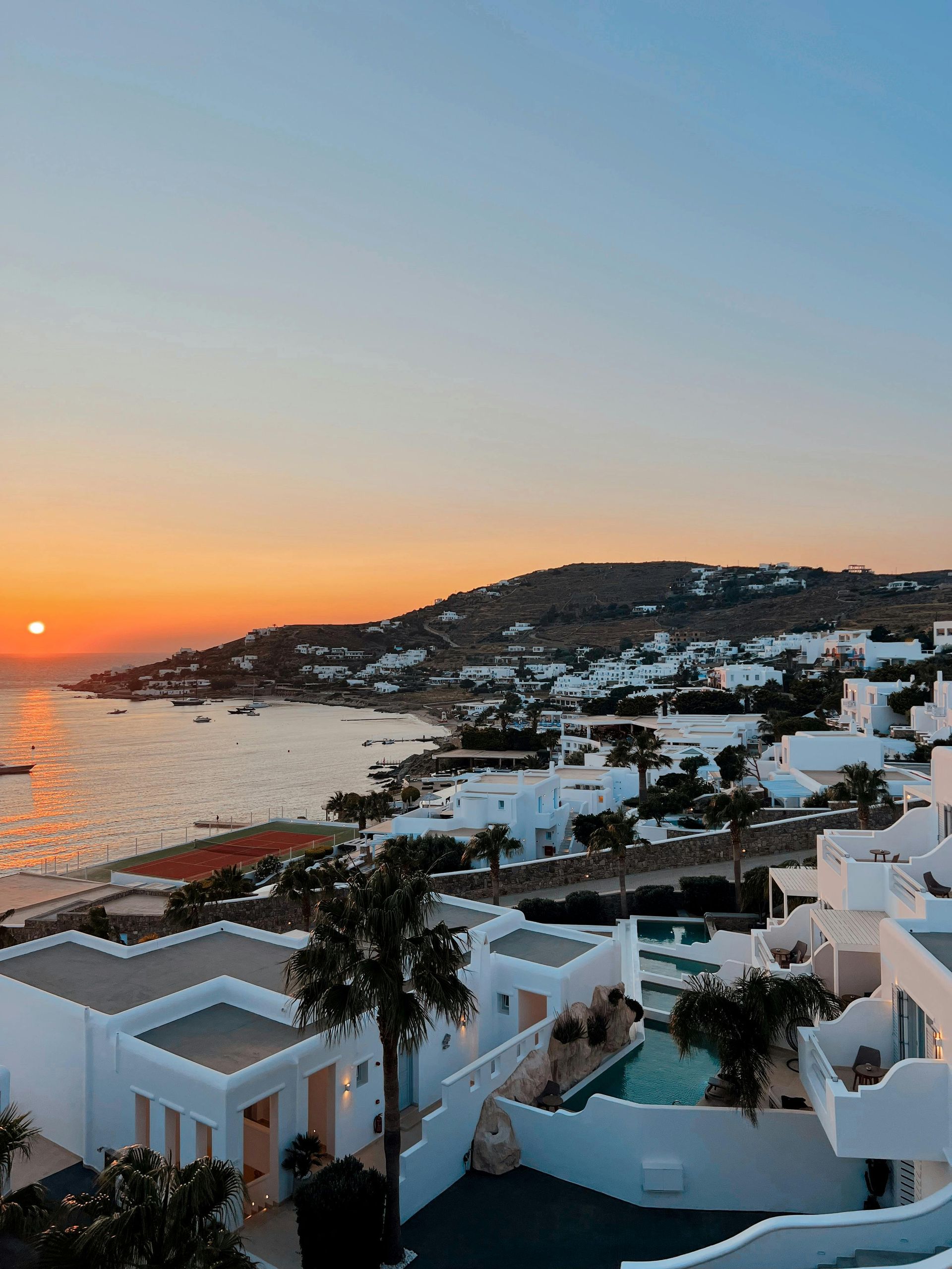 Sunset over white buildings on a coast with a palm tree, the sun reflecting on the water. Mykonos