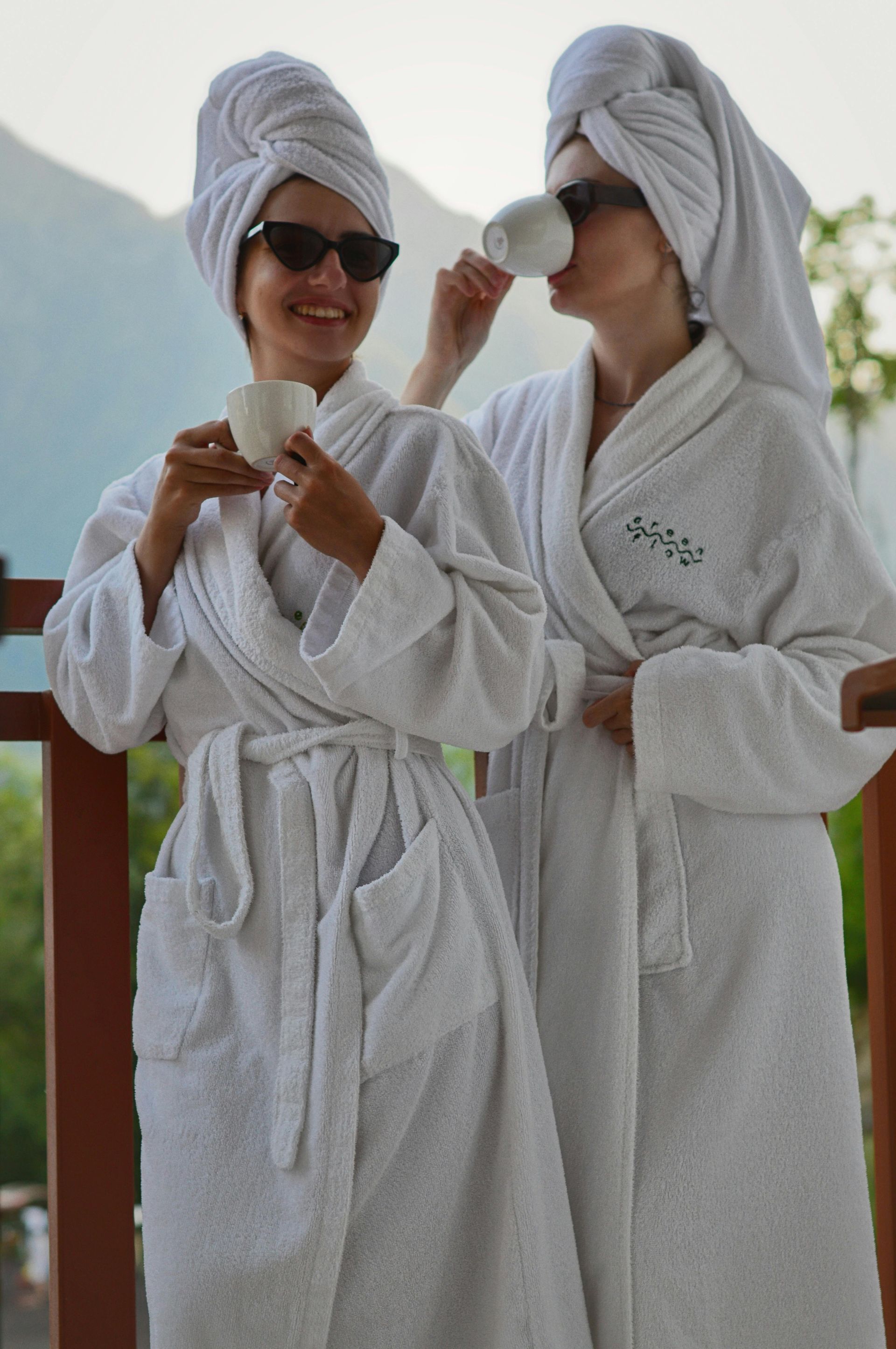 Two women in white robes and towels drinking from cups on a balcony, sunglasses on.