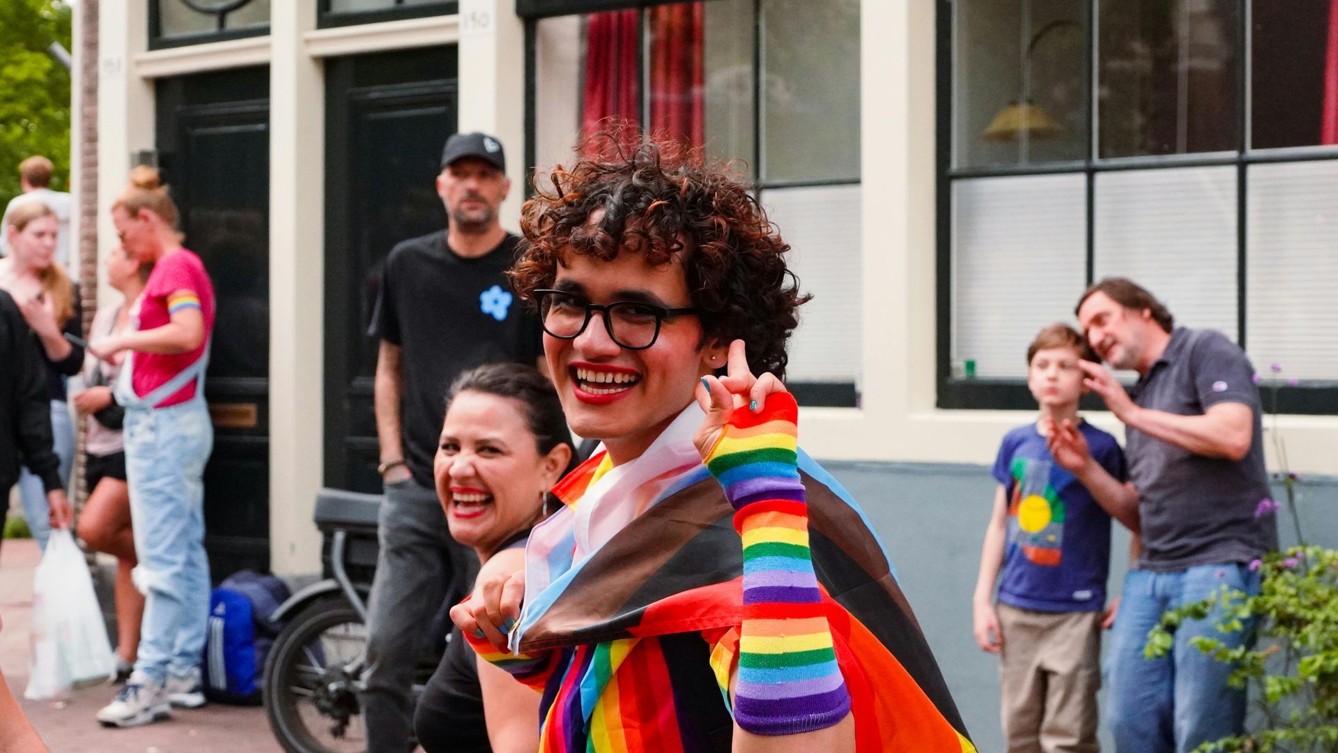 Person in rainbow gear smiles and gives a peace sign at a street event. Others watch.