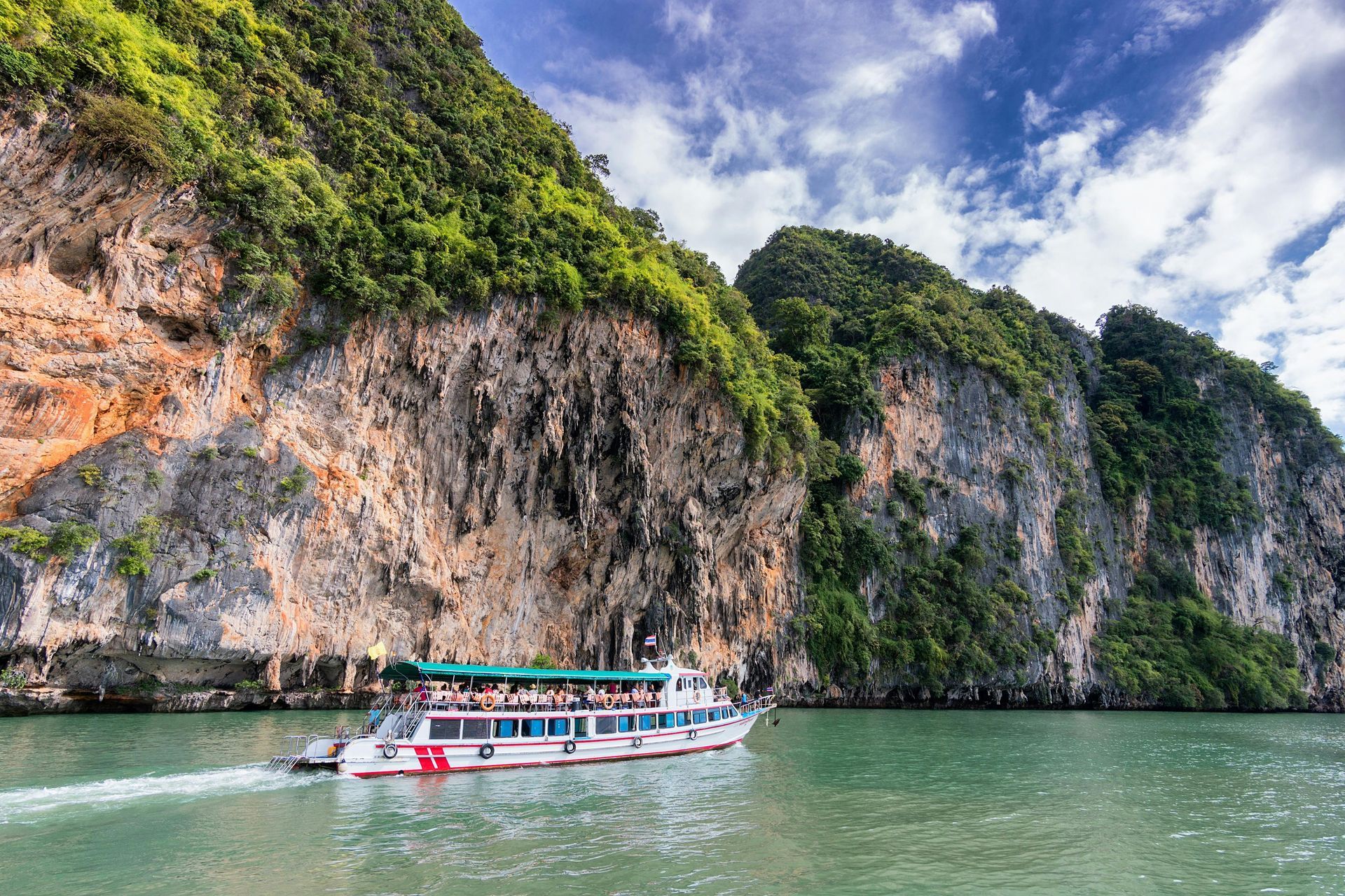 A white tourist boat sails on turquoise water near tall, green-covered cliffs under a blue sky. Thailand