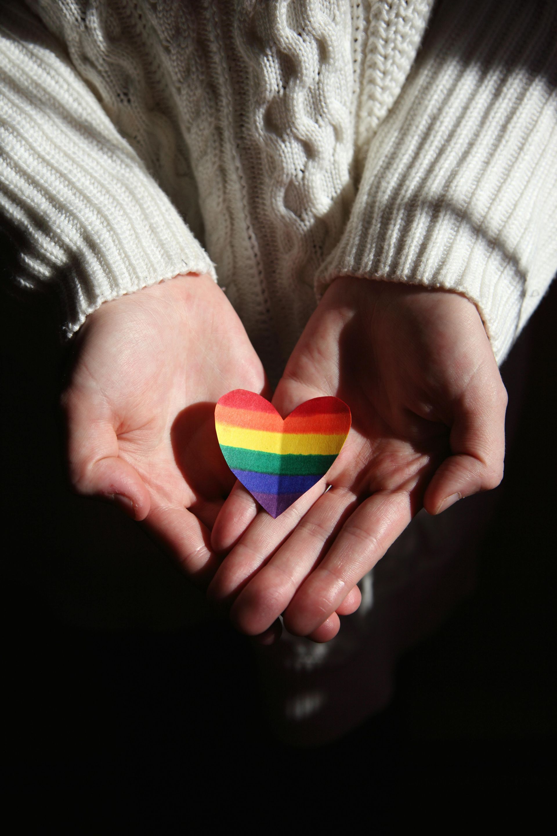 Hands cupped, holding a rainbow paper heart, person wearing a white sweater.