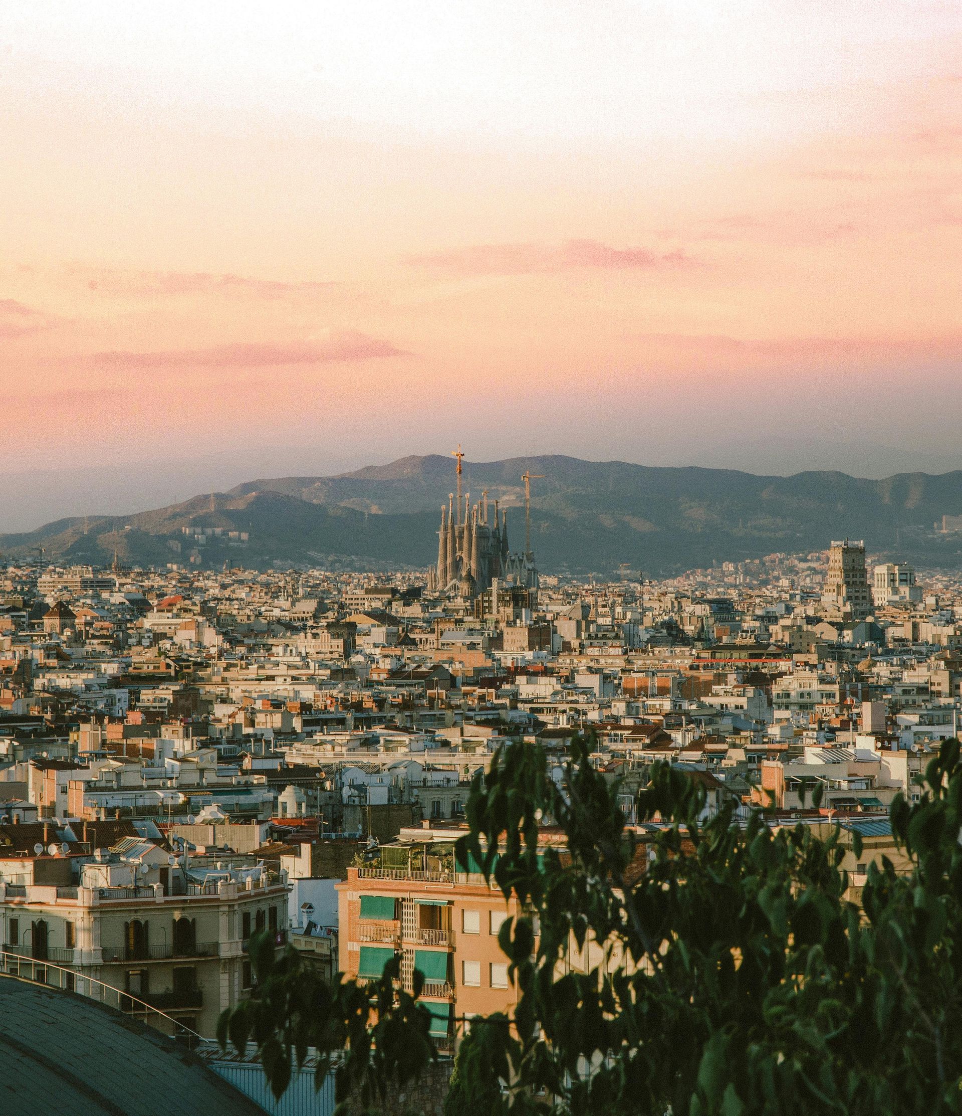 Cityscape at sunset with dense buildings, hills in the distance, and a pink-orange sky.