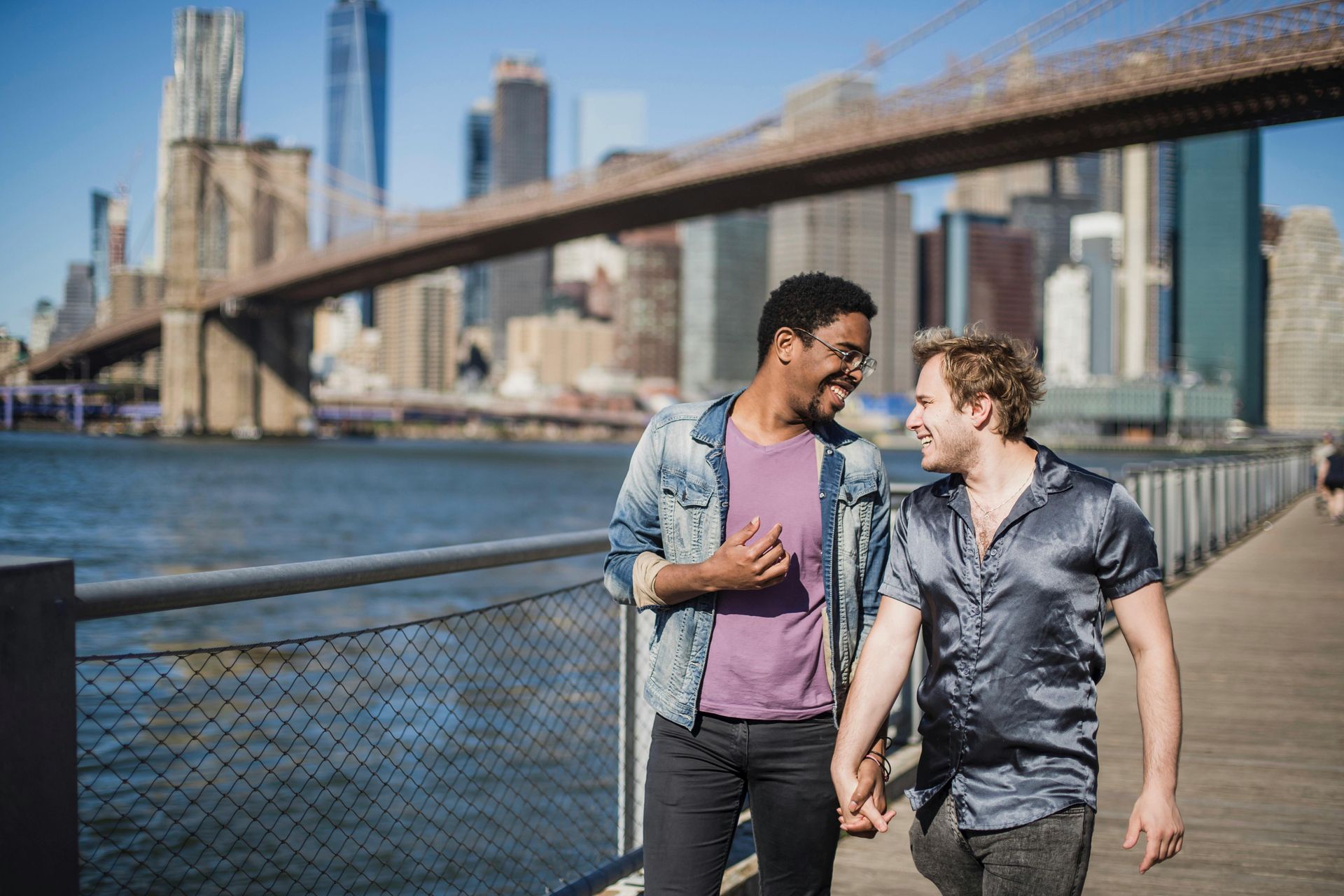 Two men holding hands, smiling, walking on a pier with Brooklyn Bridge and cityscape in the background.