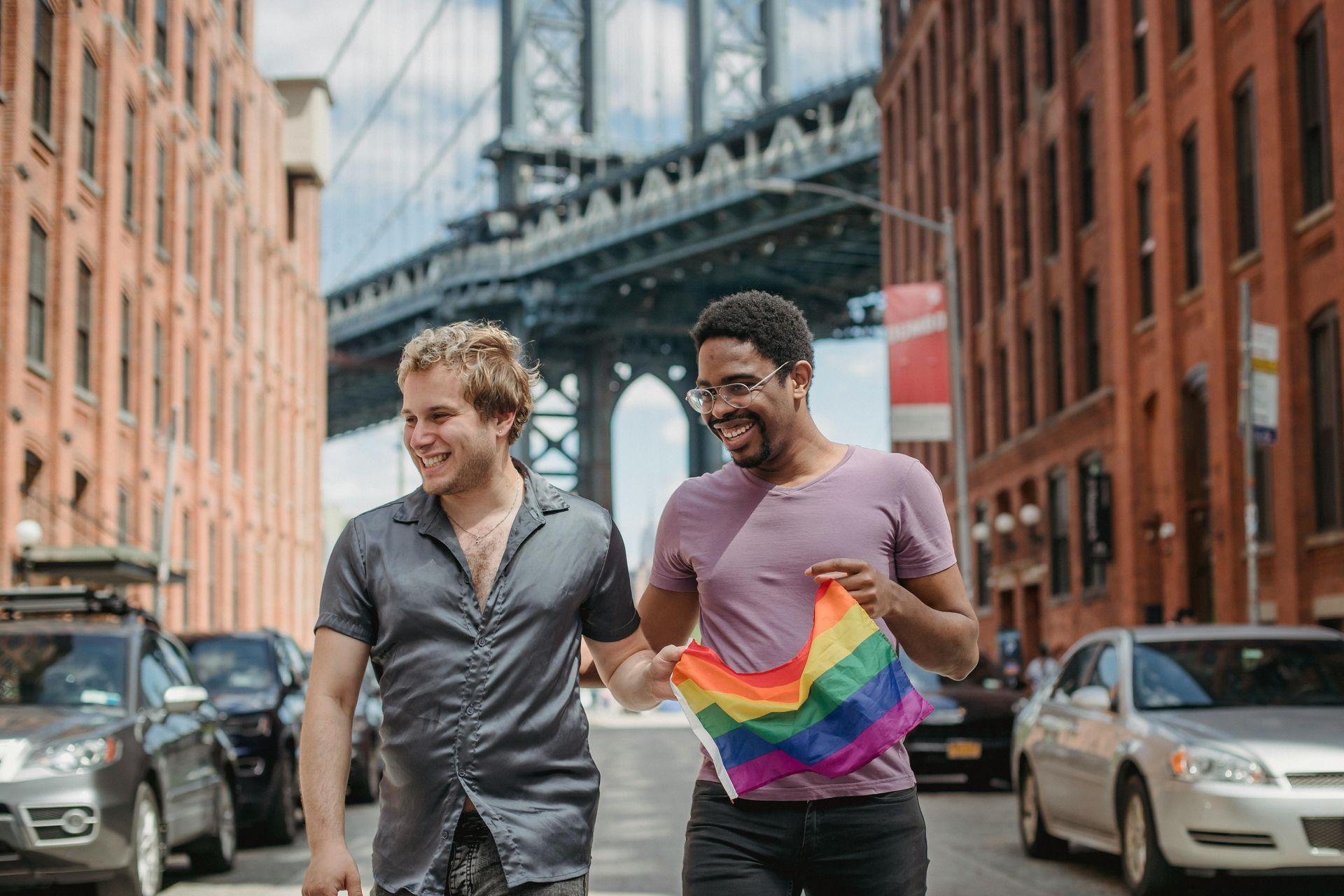 Two men walking hand-in-hand in a city street, one holding a rainbow flag. Manhattan Bridge in background.