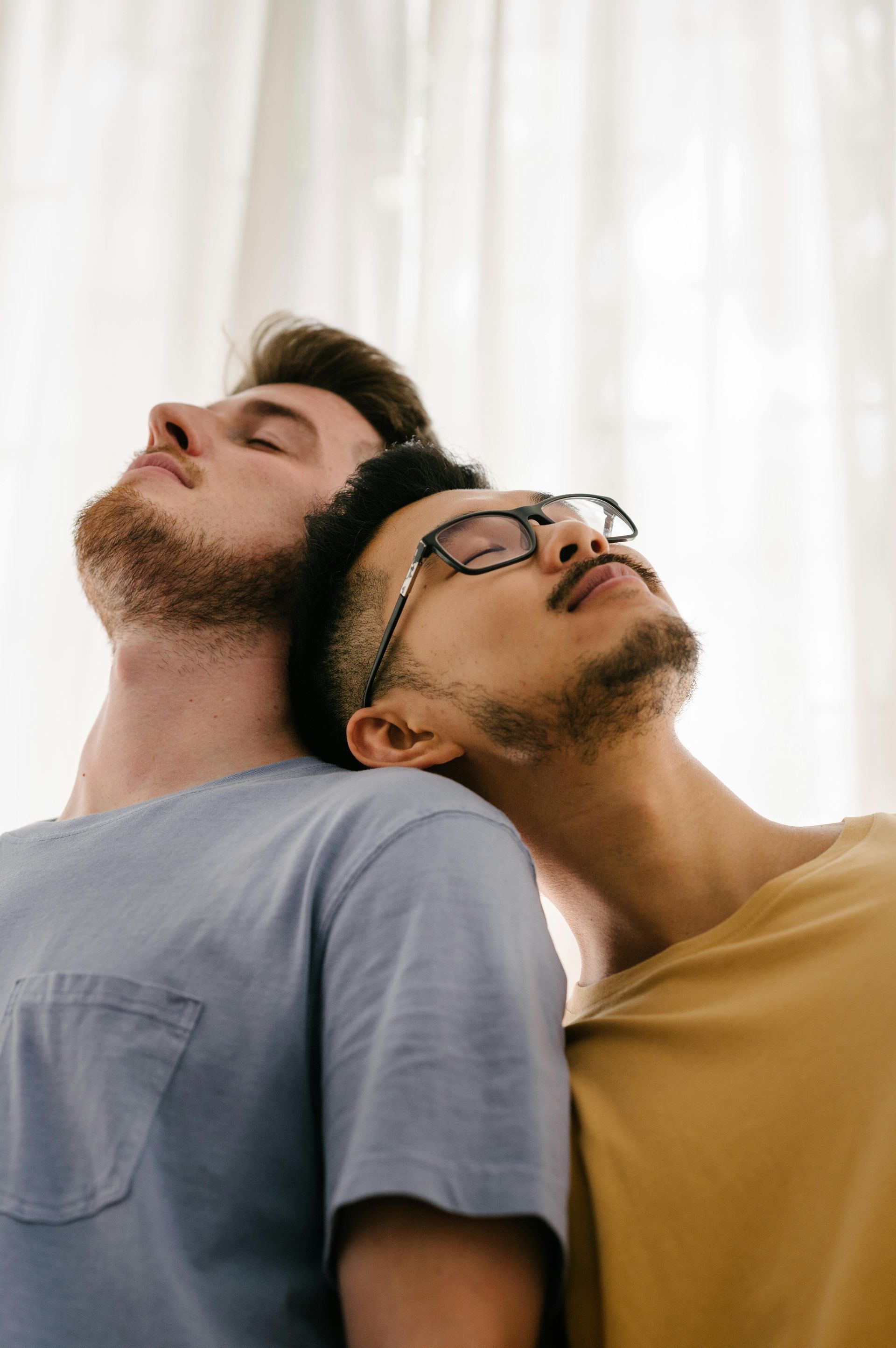 Two people with their heads tilted back, eyes closed, resting against each other in front of a bright, sheer curtain.