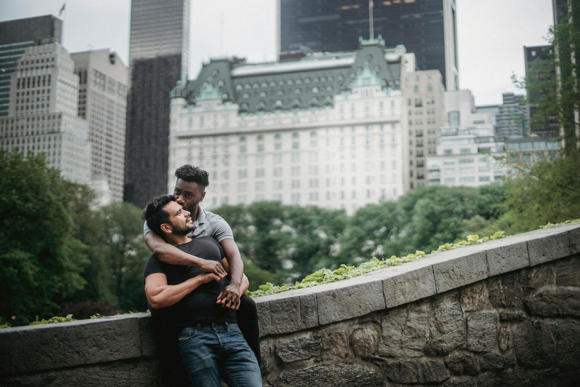 Two men embrace on a stone ledge in a city park, with buildings in the background.